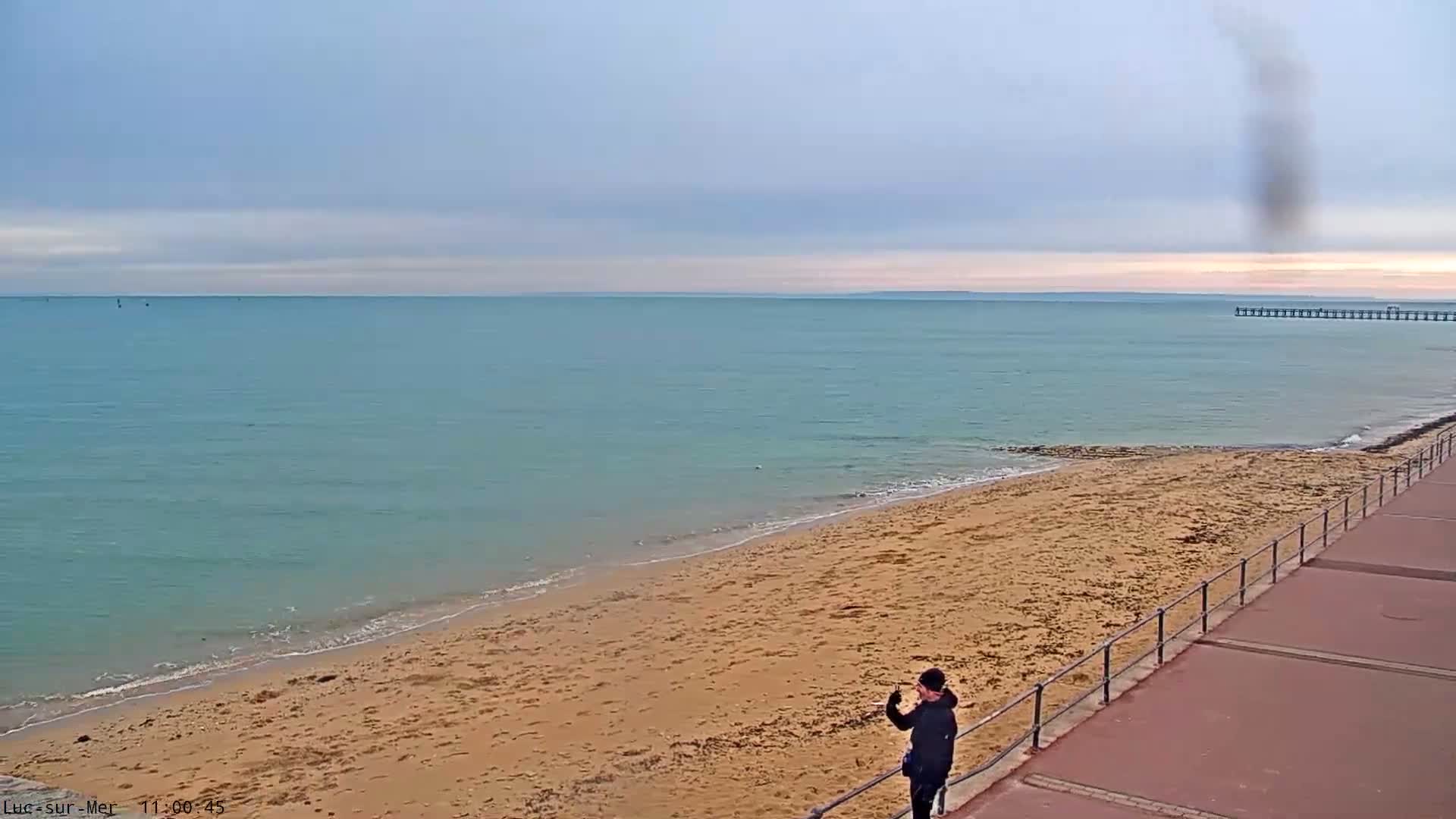 Under an overcast sky, a person stands on a seaside promenade overlooking a wide sandy beach and a calm turquoise sea with a distant pier, while a dark vertical smudge is visible in the upper right.