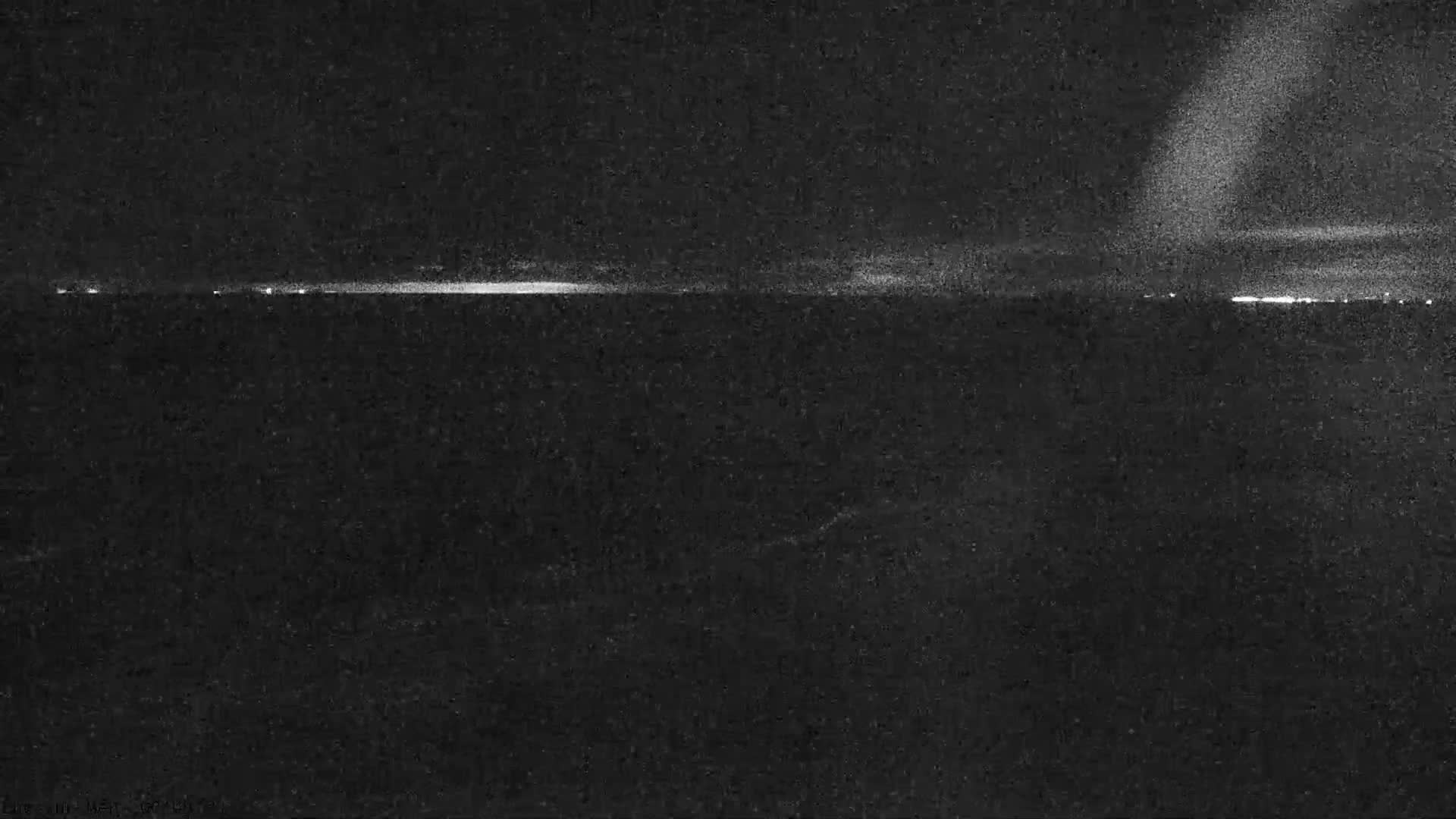 A grainy, dark night landscape shows a distant horizontal line of bright lights across the horizon, with a large, diffuse, upward-reaching glow visible in the upper right sky, suggesting hazy or partially cloudy conditions.