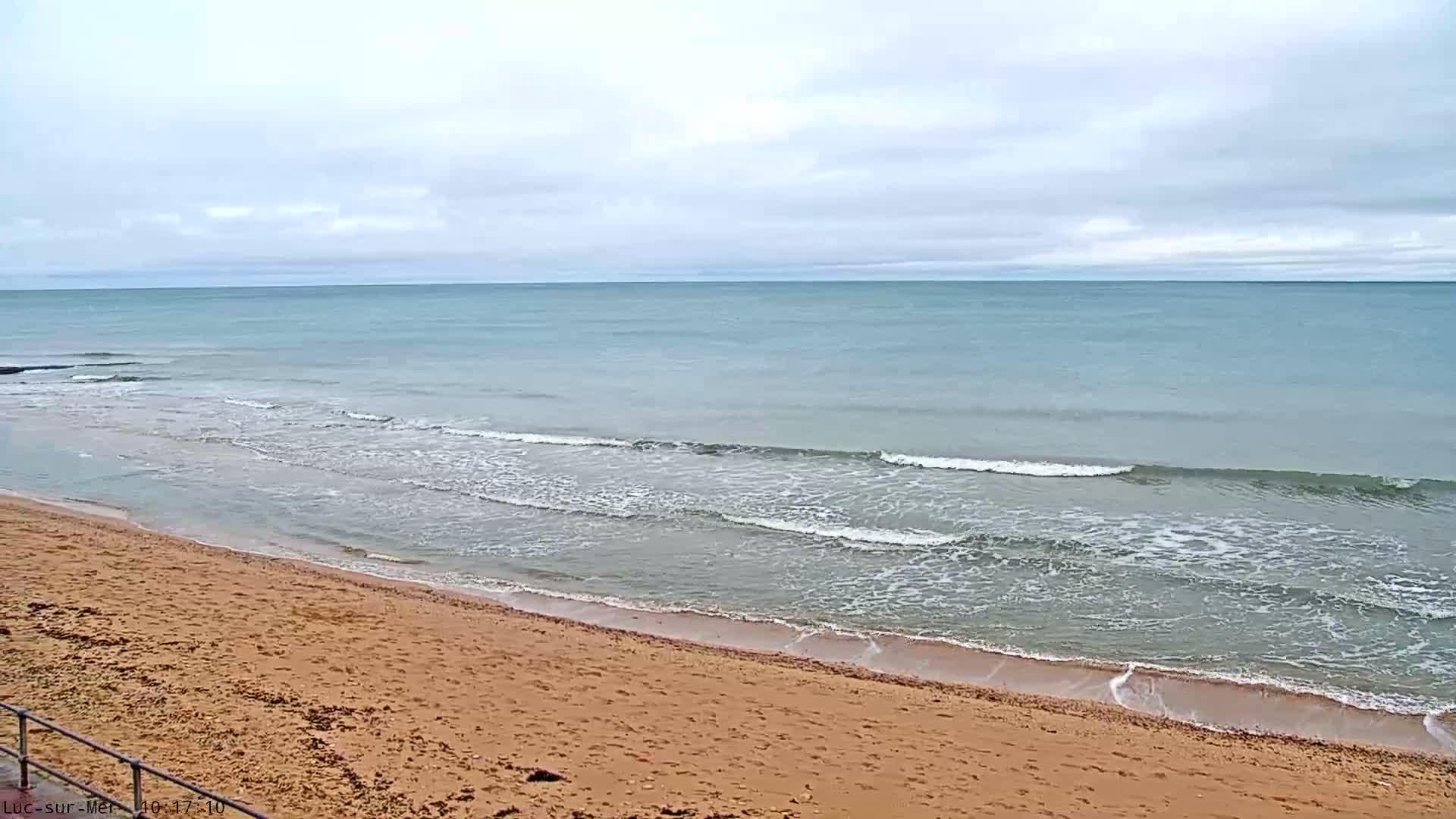 A sandy beach meets the light blue ocean with gentle waves under a gray, overcast sky.