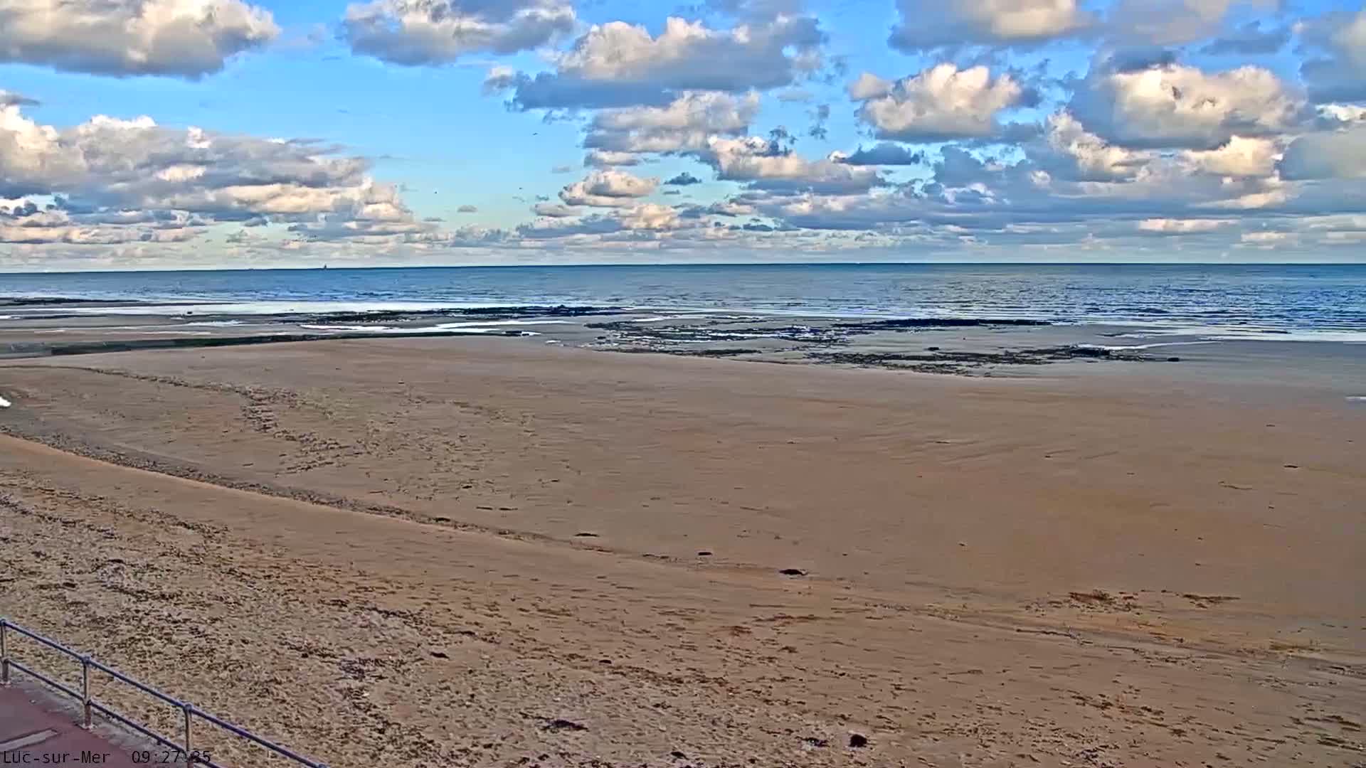 A wide sandy beach stretches towards the calm blue ocean under a partly cloudy sky with scattered white clouds.