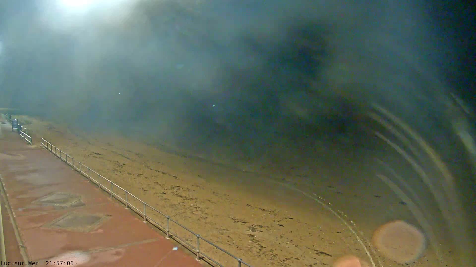 A dark and extremely foggy outdoor scene shows a wet reddish promenade with railings bordering a sandy beach, with the obscured sea creating extremely poor visibility.