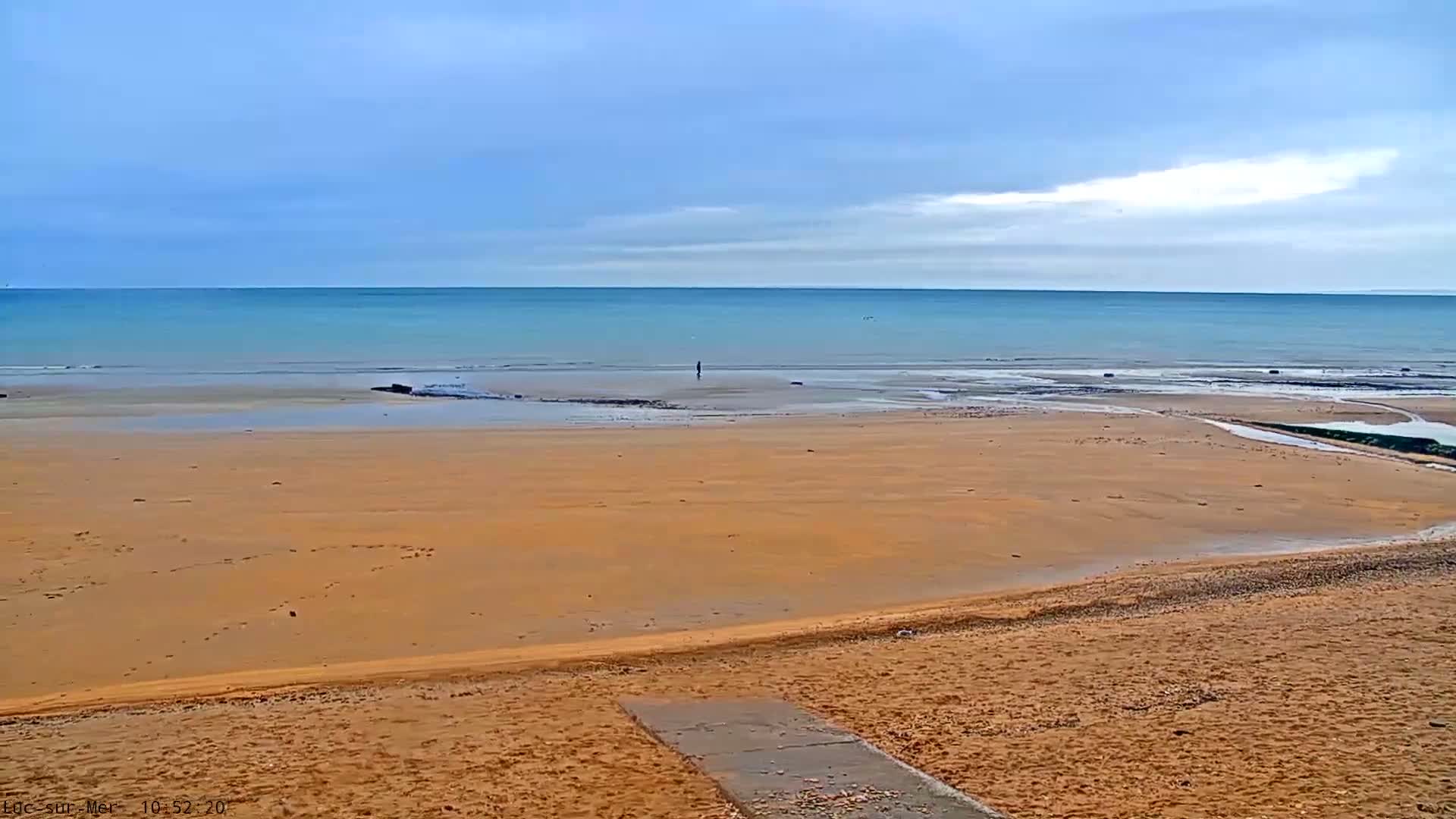 A wide, golden-brown sandy beach features a small stream flowing into the calm, blue-green sea where a lone figure stands, all beneath a cloudy sky.