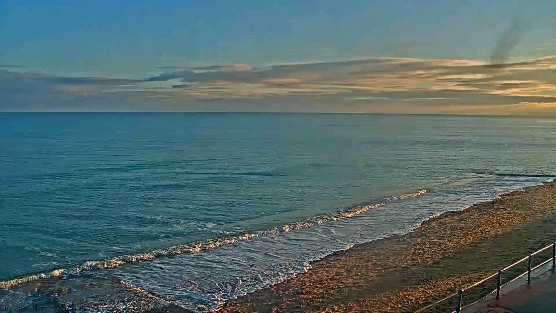 A tranquil scene shows a calm blue-green ocean with gentle waves breaking on a sandy beach, viewed under a partly cloudy sky with golden light on the horizon, with a metal railing visible in the foreground.