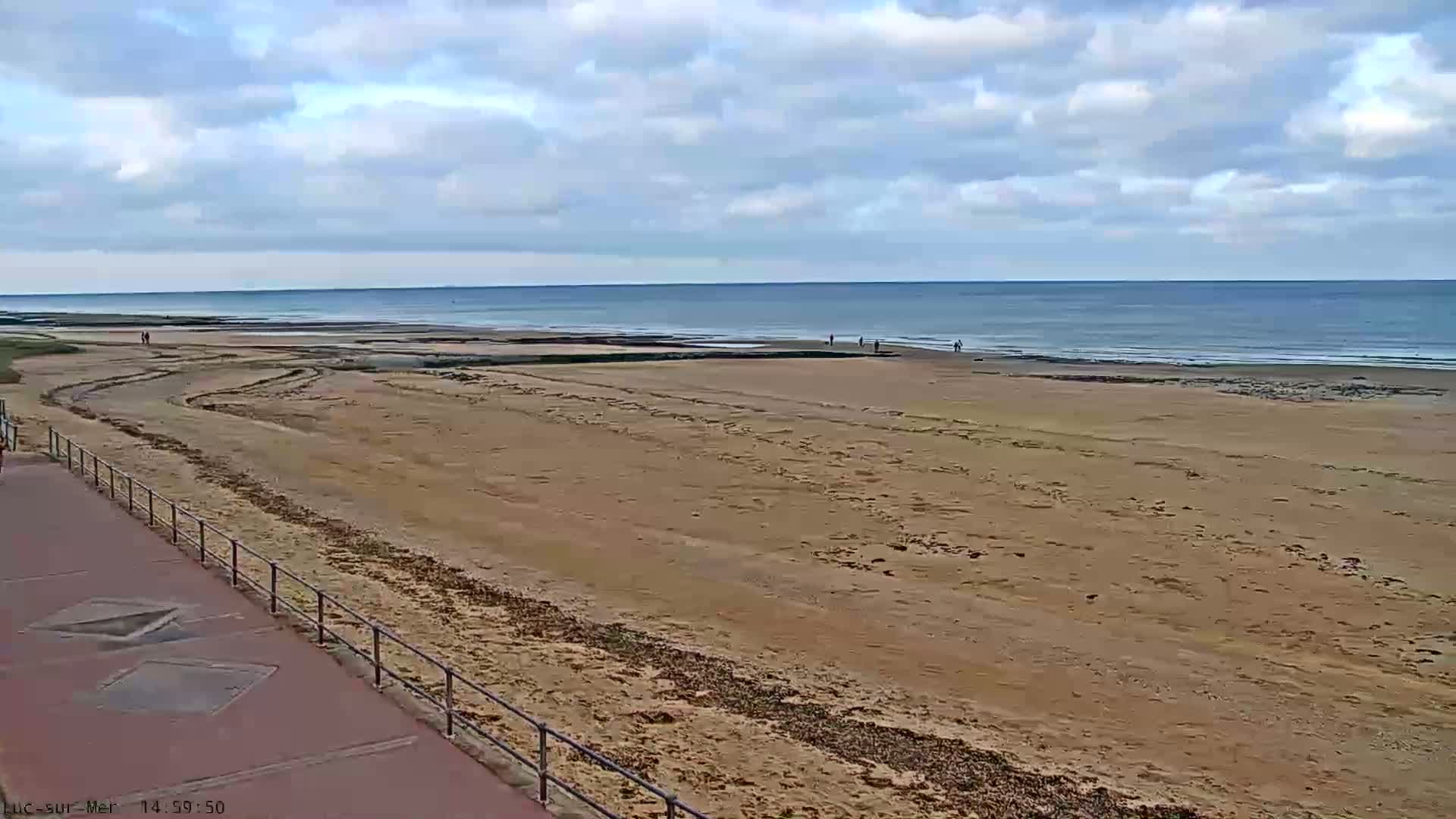 A tranquil scene shows a calm blue-green ocean with gentle waves breaking on a sandy beach, viewed under a partly cloudy sky with golden light on the horizon, with a metal railing visible in the foreground.
