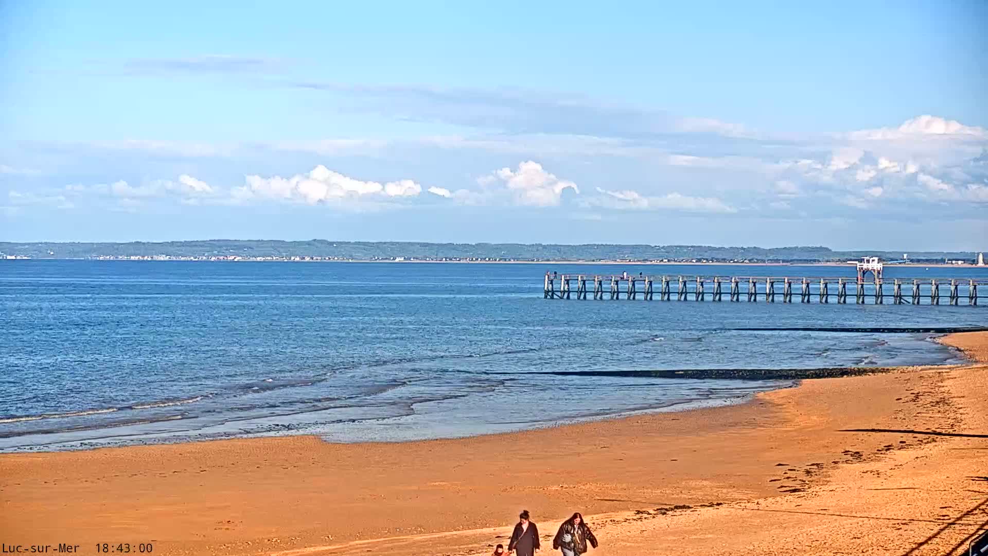 A tranquil scene shows a calm blue-green ocean with gentle waves breaking on a sandy beach, viewed under a partly cloudy sky with golden light on the horizon, with a metal railing visible in the foreground.