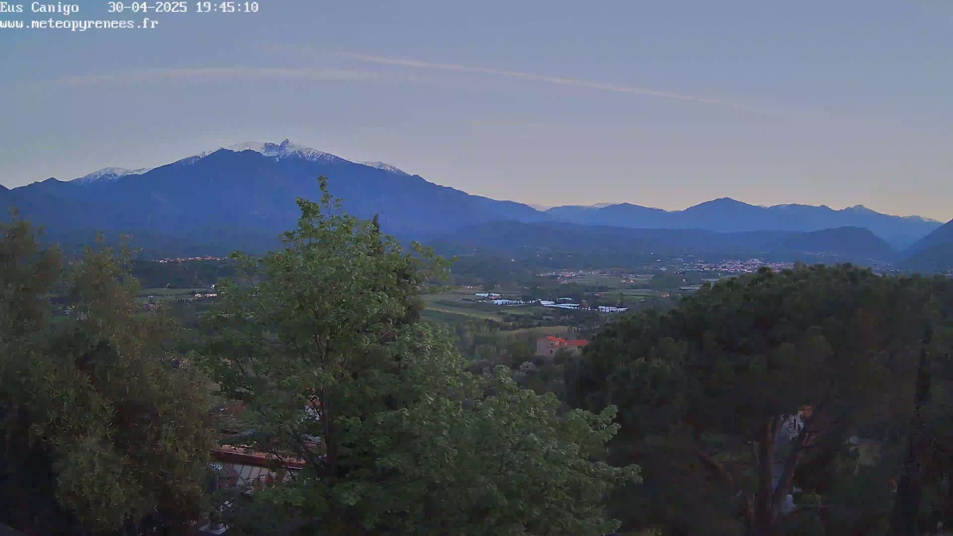 A mountain range with snow-capped peaks is visible in the distance, overlooking a valley with trees and a town under a twilight sky.