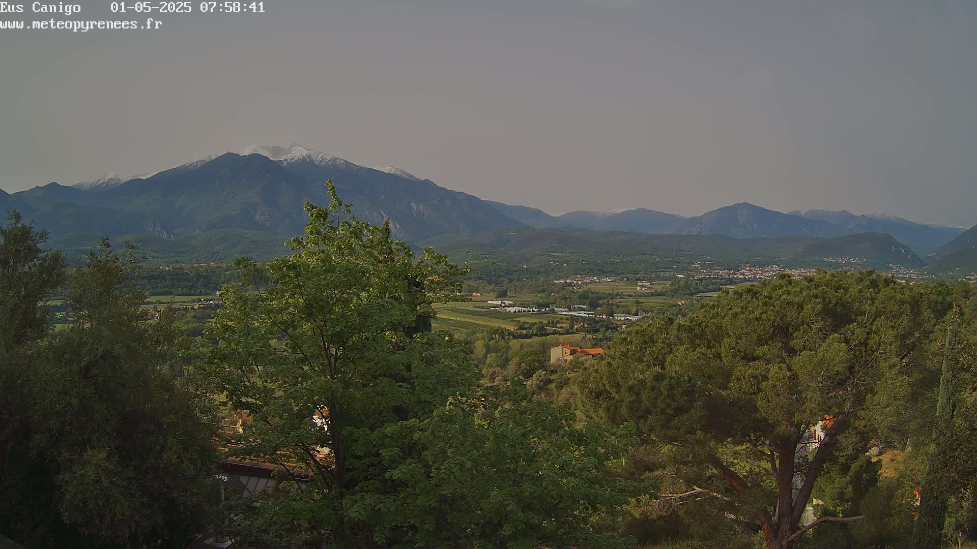 A view from a hilltop shows a valley with green trees and buildings, with mountains in the background, one of which has snow on its peak, under a hazy sky.