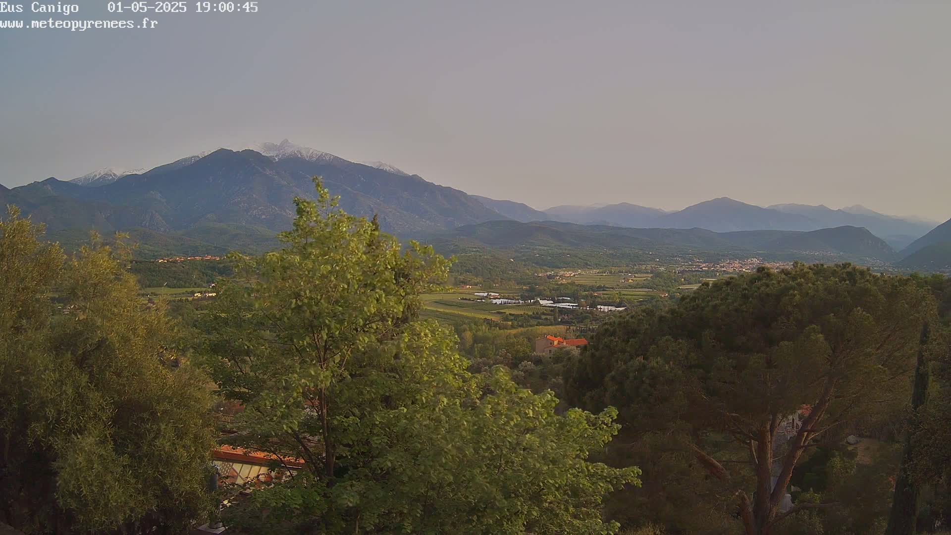 A panoramic view shows a valley with greenery and a small town nestled between mountains, one of which is snow-capped, under a pale, clear sky.