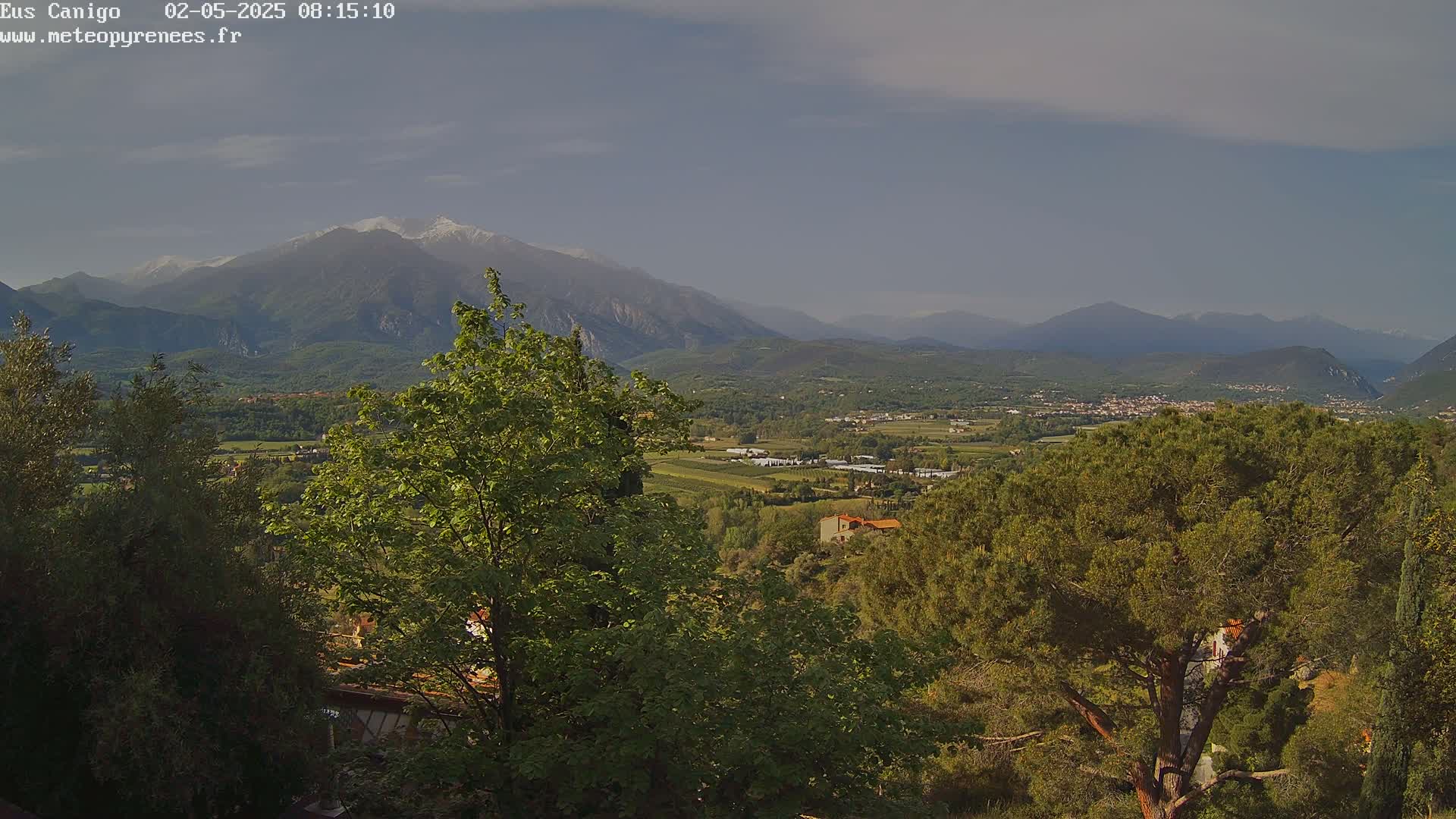 A partly cloudy day reveals a snow-capped mountain range in the distance, overlooking a valley of green trees, shrubs, and buildings.