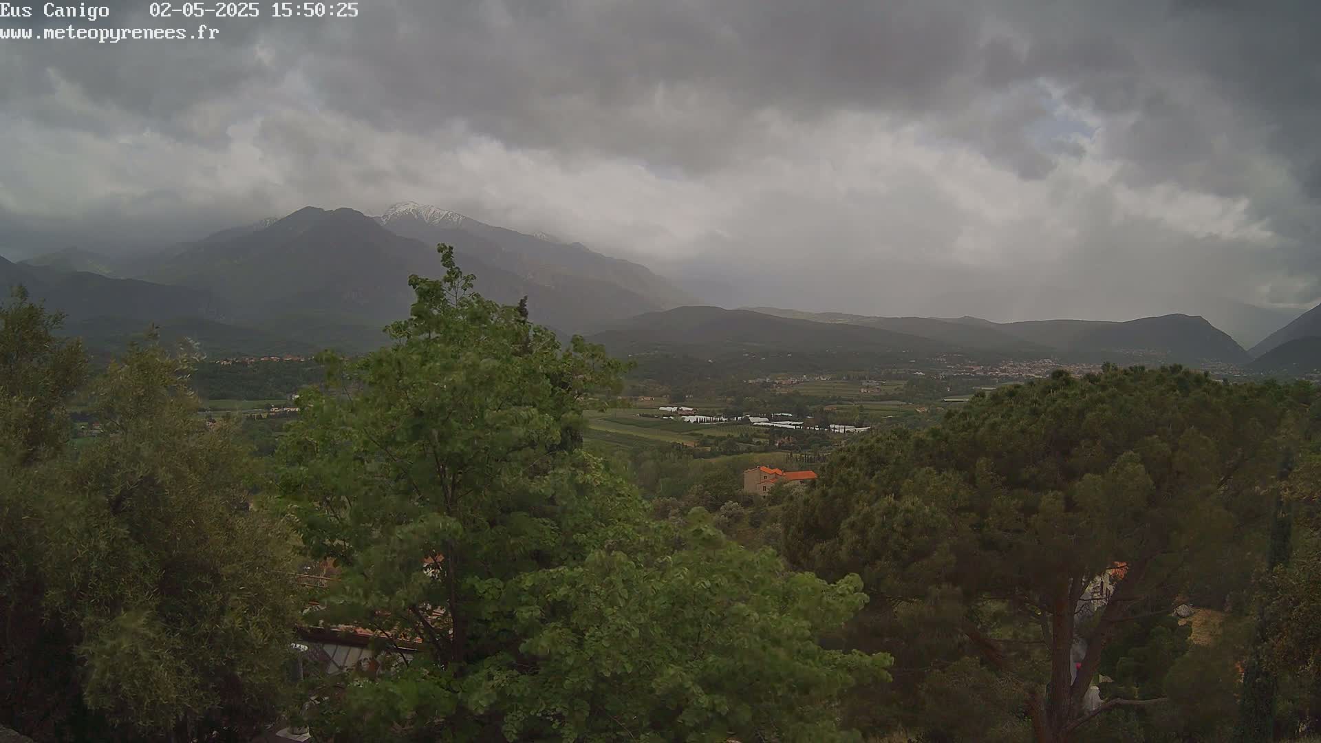A view of a valley dotted with trees and buildings under a cloudy, overcast sky, with snow-capped mountains in the background.