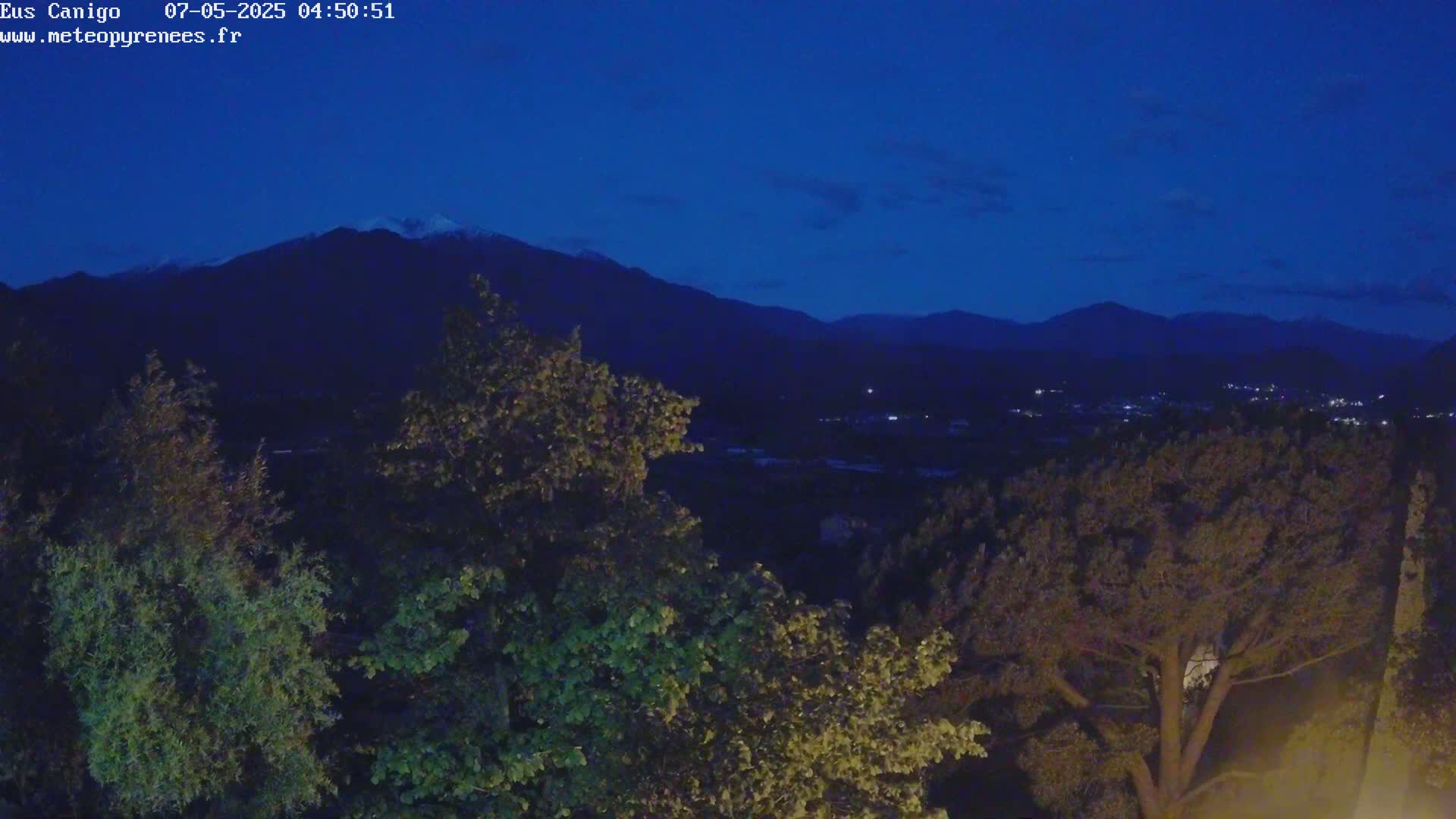 A dark blue night sky overlooks a valley of dark green trees and distant mountains, with one mountain peak showing a hint of snow.