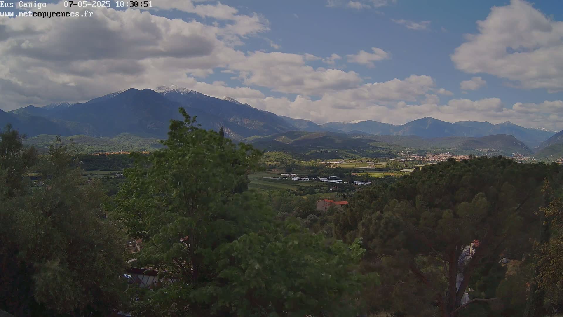 A partly cloudy sky overlooks a valley of green trees and fields, with snow-capped mountains in the distance.