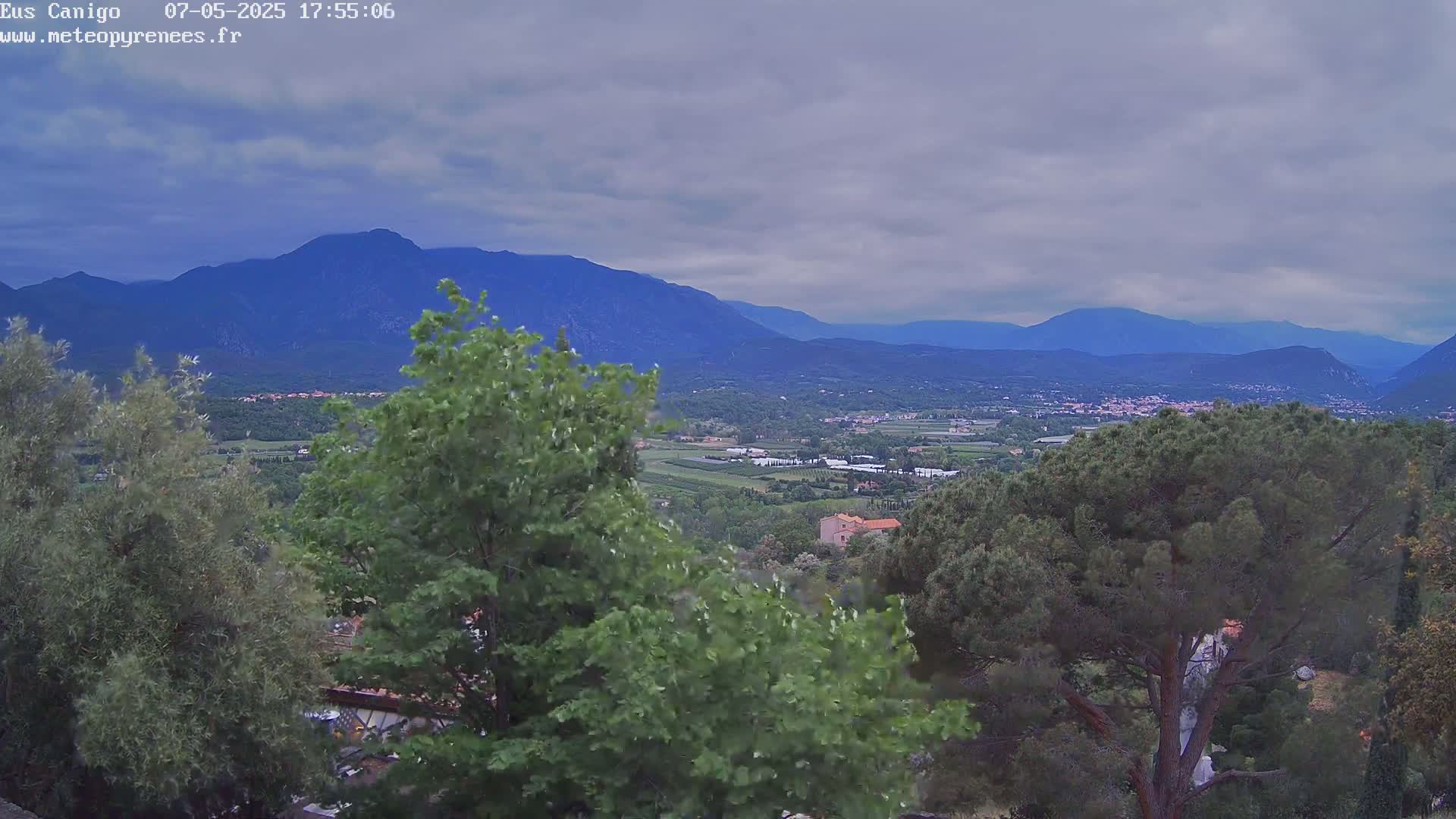 A valley dotted with trees and buildings stretches out beneath a cloudy sky, backed by a range of blue mountains.