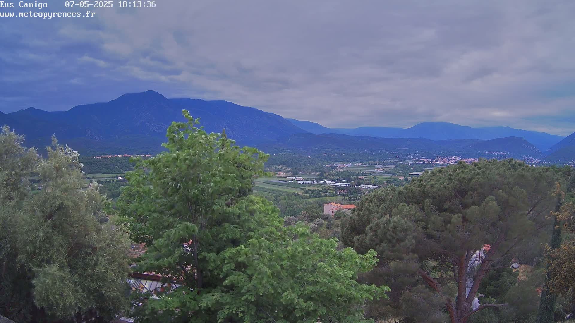 A mountainous landscape under a cloudy sky is seen from an elevated vantage point, showing a valley with greenery and a village in the distance.