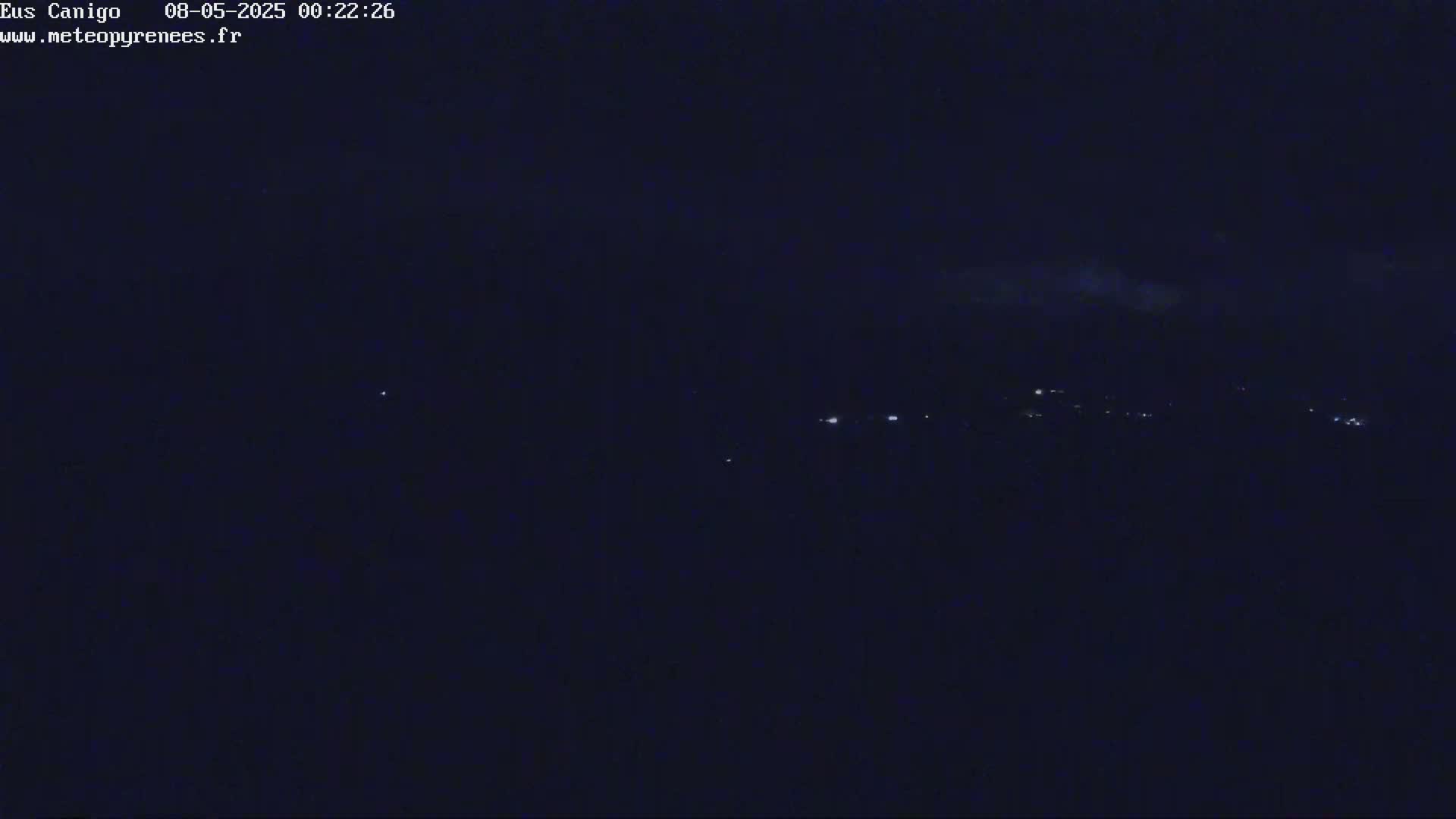 A dark nighttime scene shows a few distant lights on a hill under a mostly clear, dark sky.