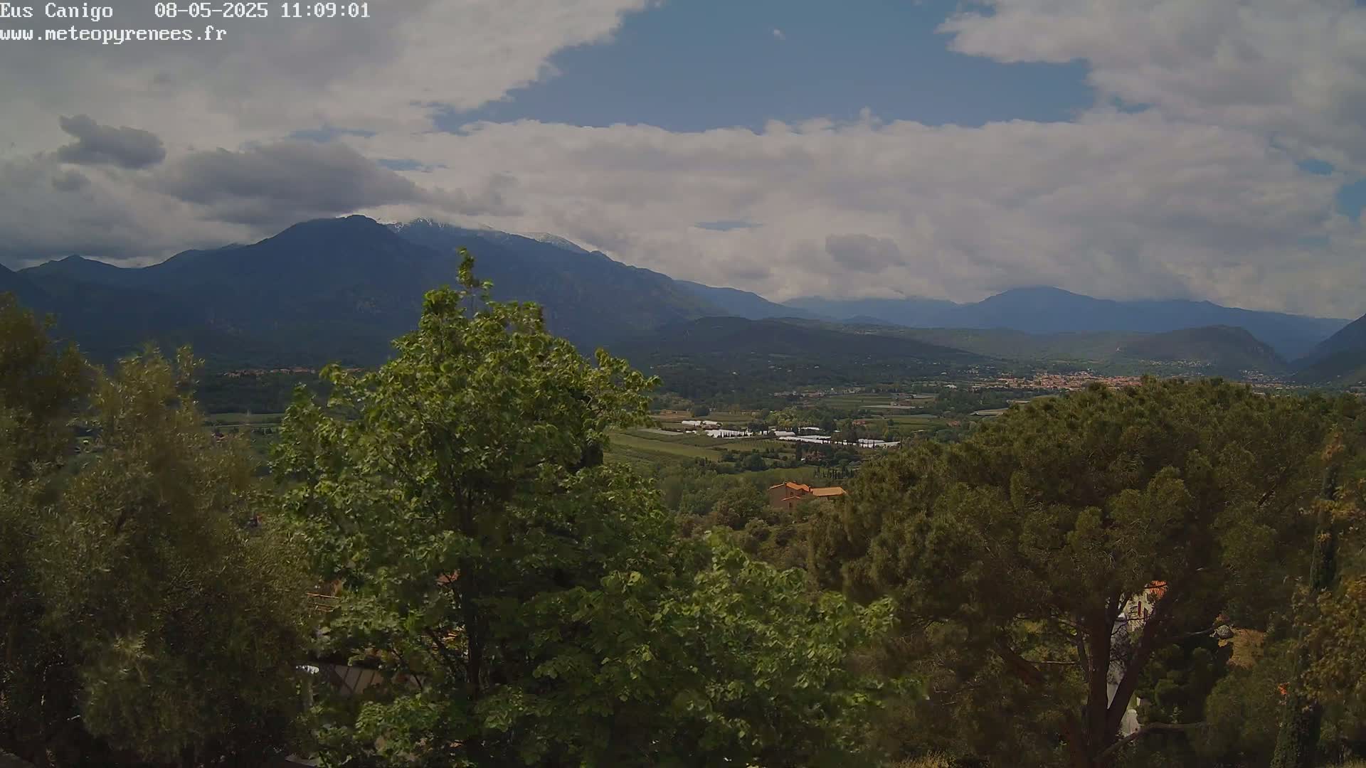 A partly cloudy day reveals a valley with green trees and distant mountains, some with snow-capped peaks.