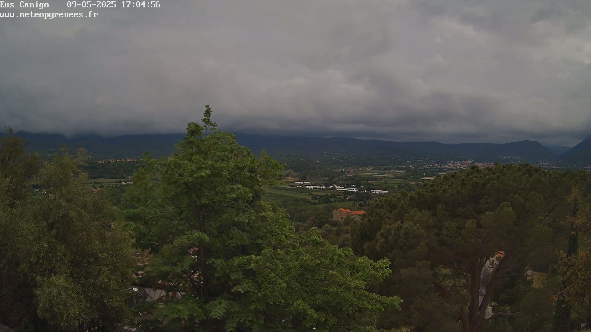 A view from a hilltop shows a valley with trees, buildings, and farmland under a heavily clouded sky.