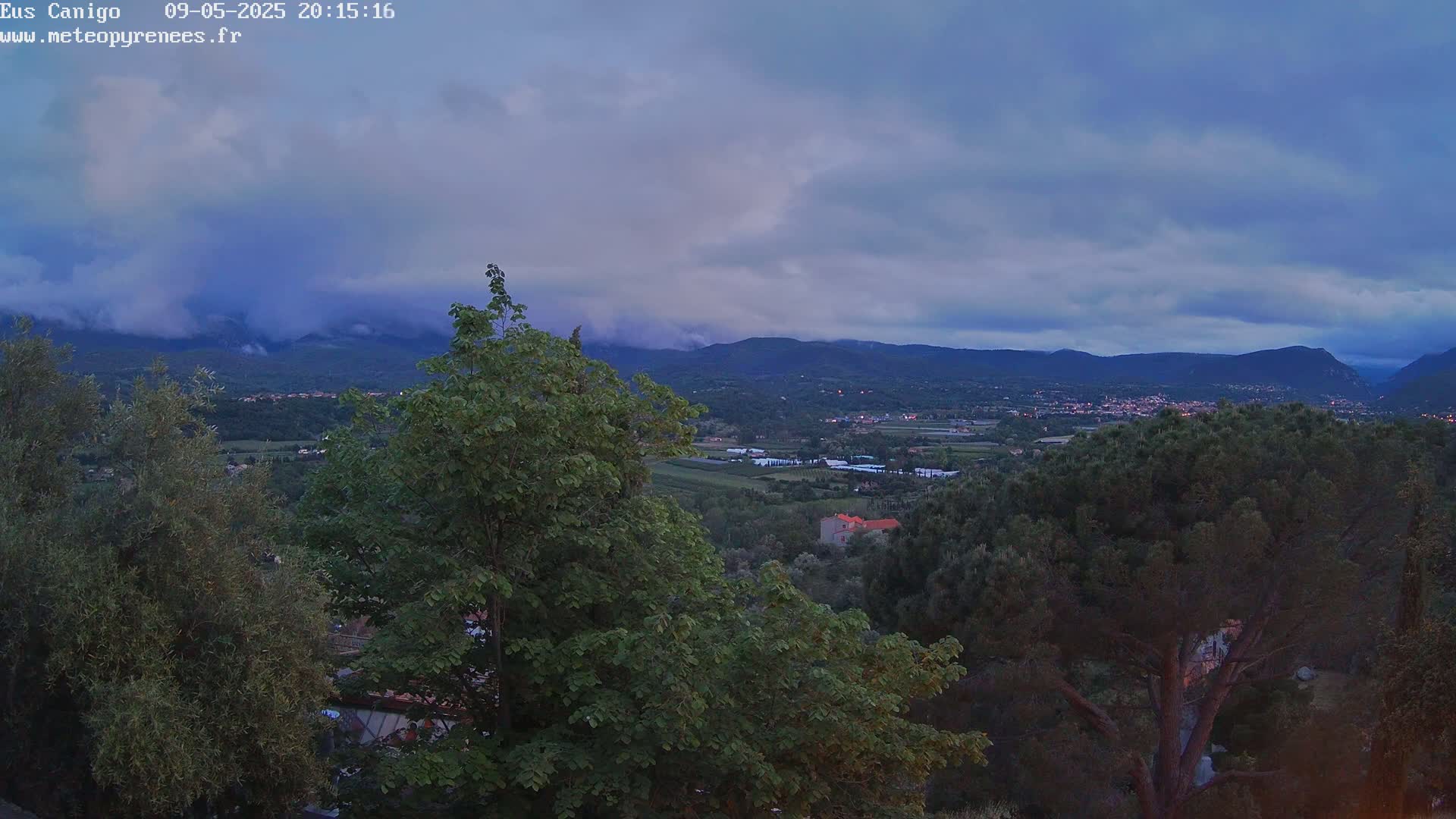 A view of a valley at dusk, with lush green trees in the foreground and mountains in the background under a cloudy sky.