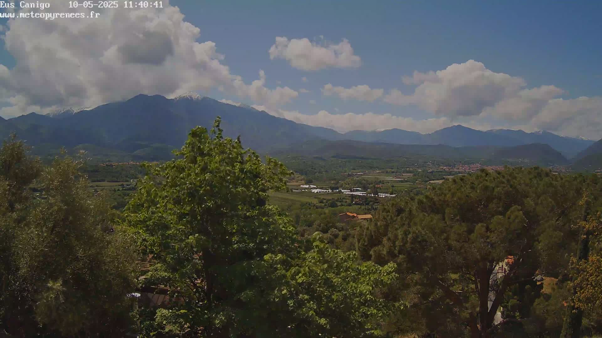 A partly cloudy day reveals a valley landscape with lush green trees and mountains in the distance, some with snow-capped peaks.