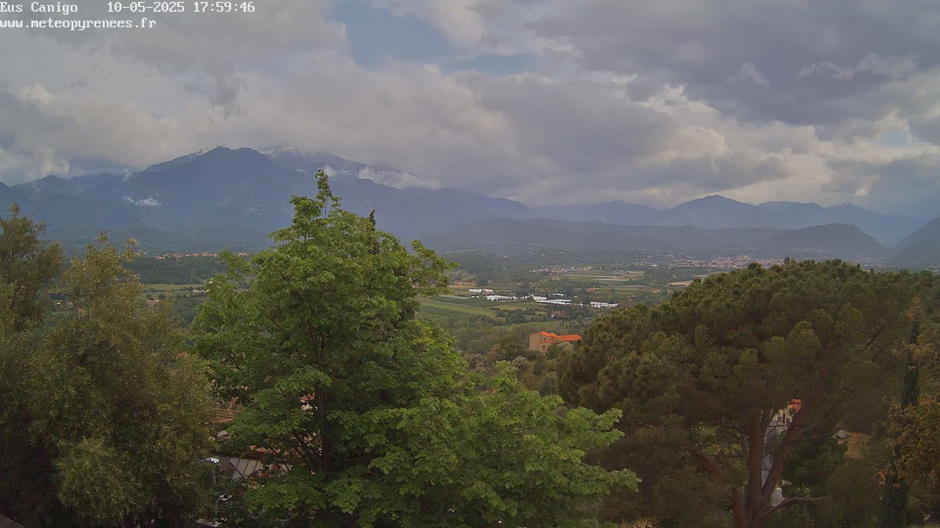 A mountain range with snow-capped peaks is visible in the distance under a cloudy sky, beyond a valley of greenery and trees.