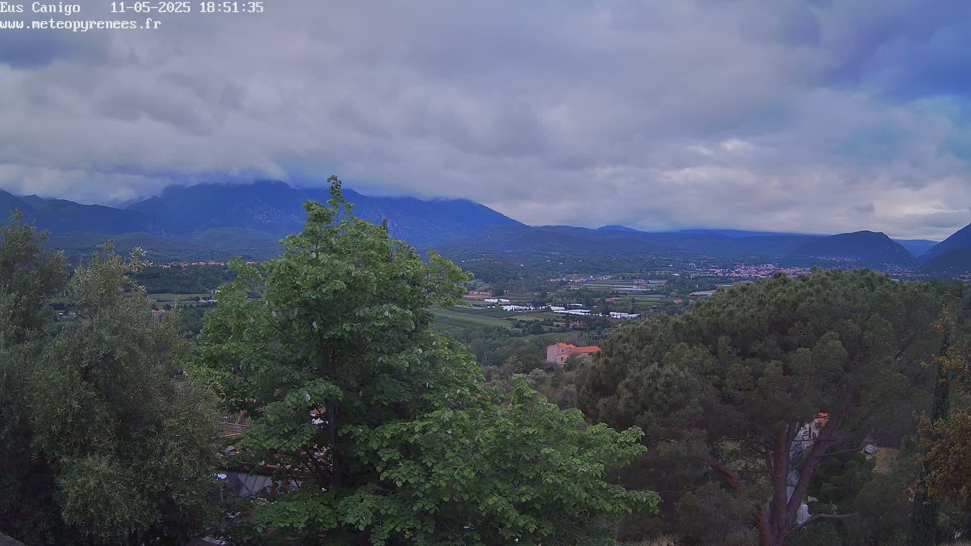 A mountainous landscape under a cloudy sky is visible from a vantage point among green trees.