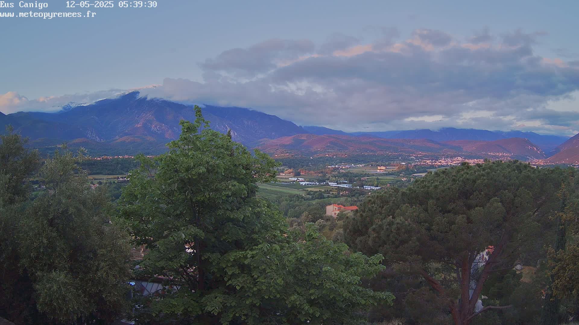 A mountain range with snow-capped peaks is visible in the distance under a partly cloudy sky, with a valley and green foliage in the foreground.