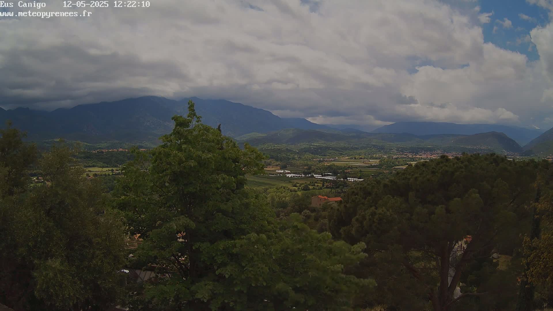 A partly cloudy sky overlooks a valley of green trees, fields, and small buildings, with mountains in the background.