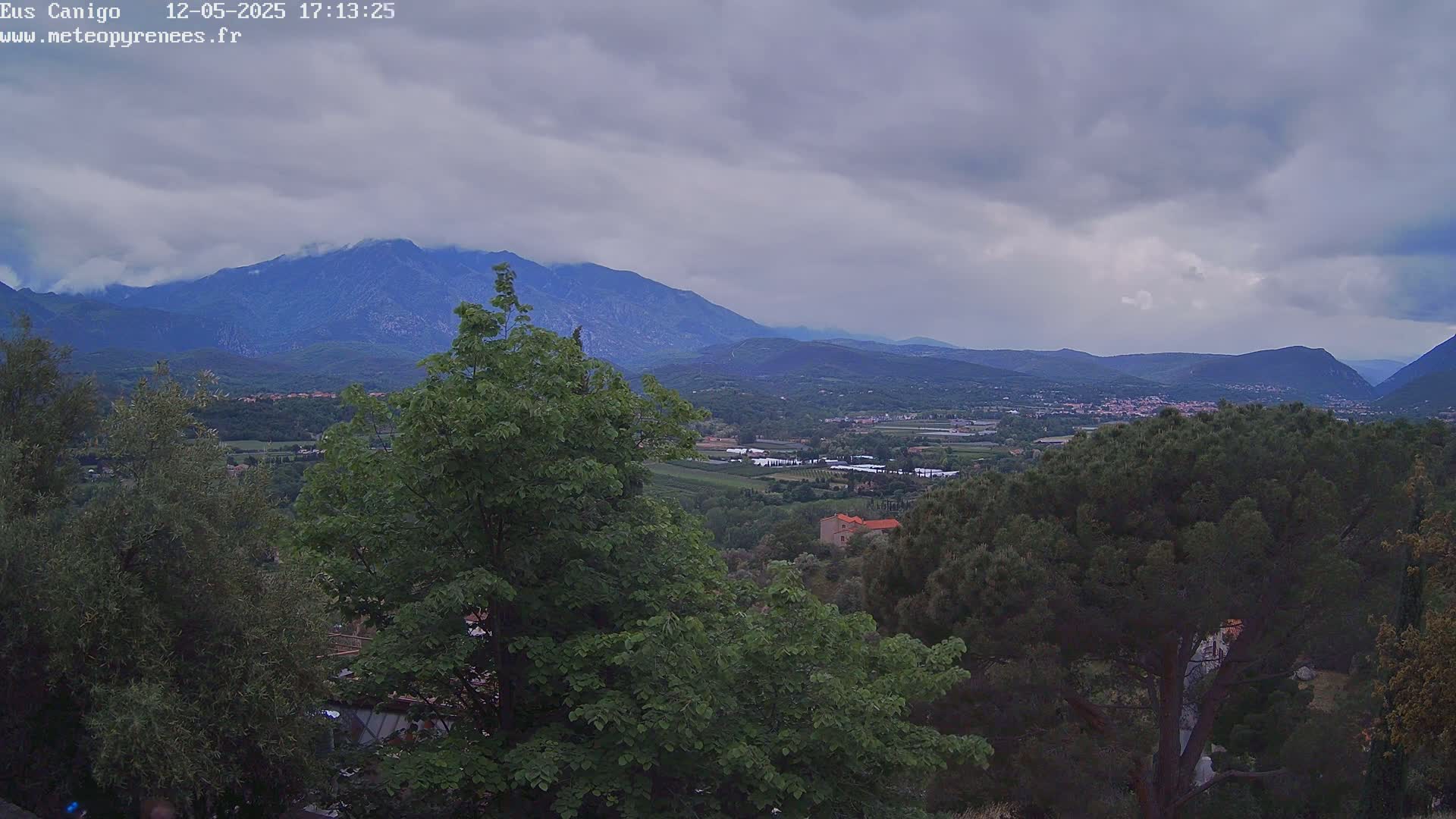 A mountain range is visible in the distance under a cloudy sky, with green trees and a small town nestled in the valley below.