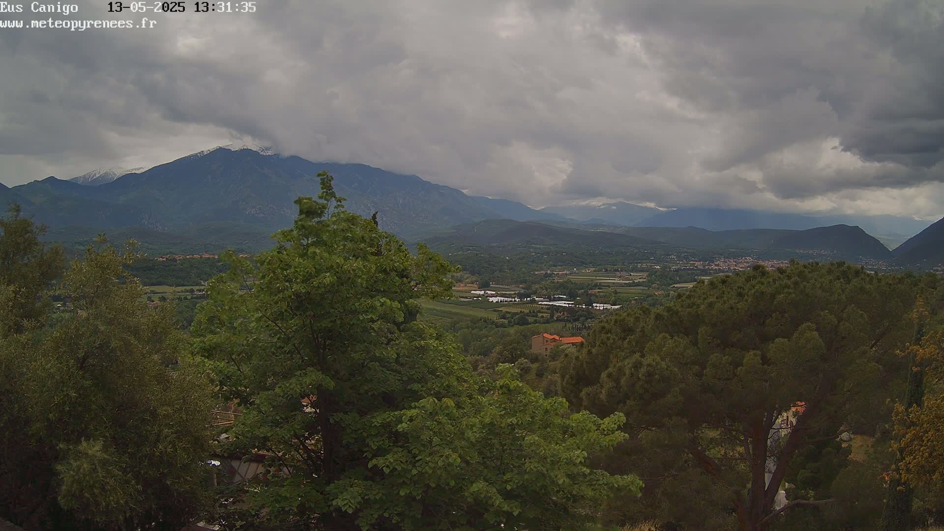A mountainous landscape under a cloudy sky is partially obscured by lush green trees in the foreground.