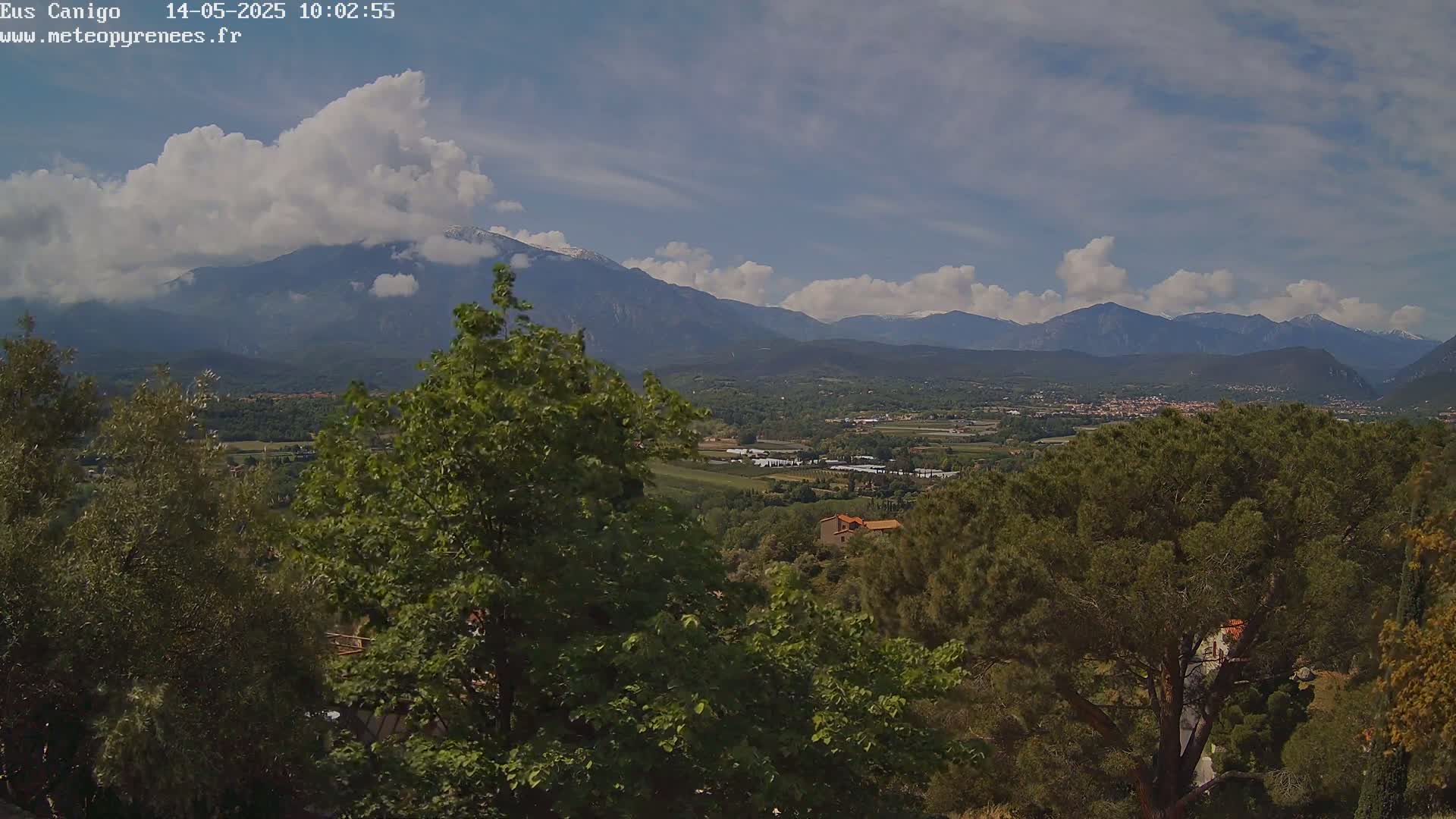 A partly cloudy day reveals a mountainous landscape with a valley containing buildings and trees in the foreground.