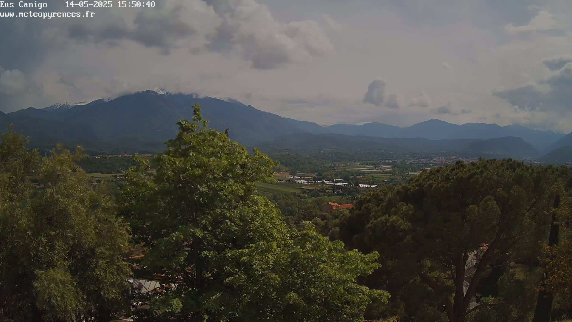 A partly cloudy day reveals a valley dotted with trees and buildings, stretching towards distant mountains with snow-capped peaks.