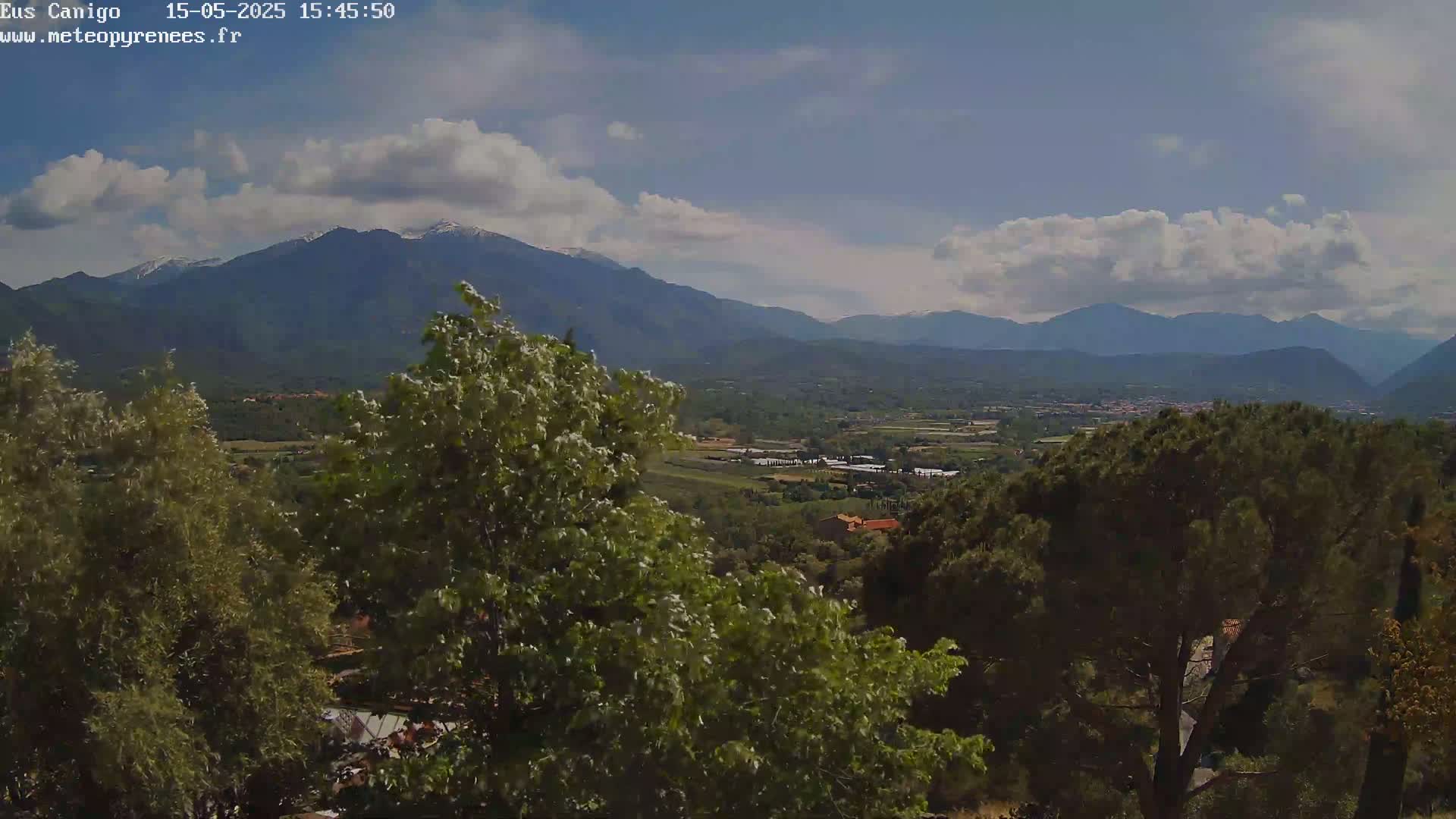 A partly cloudy day reveals a snow-capped mountain range in the distance, overlooking a valley of green trees and a small town.