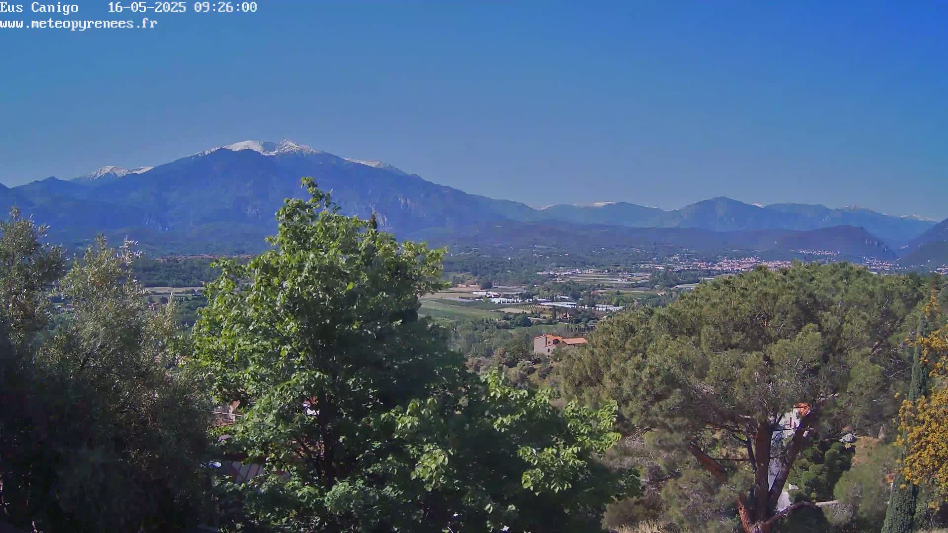 A long-range view shows snow-capped mountains in the distance under a clear blue sky, with a valley of green trees and buildings in the midground and more trees in the foreground.