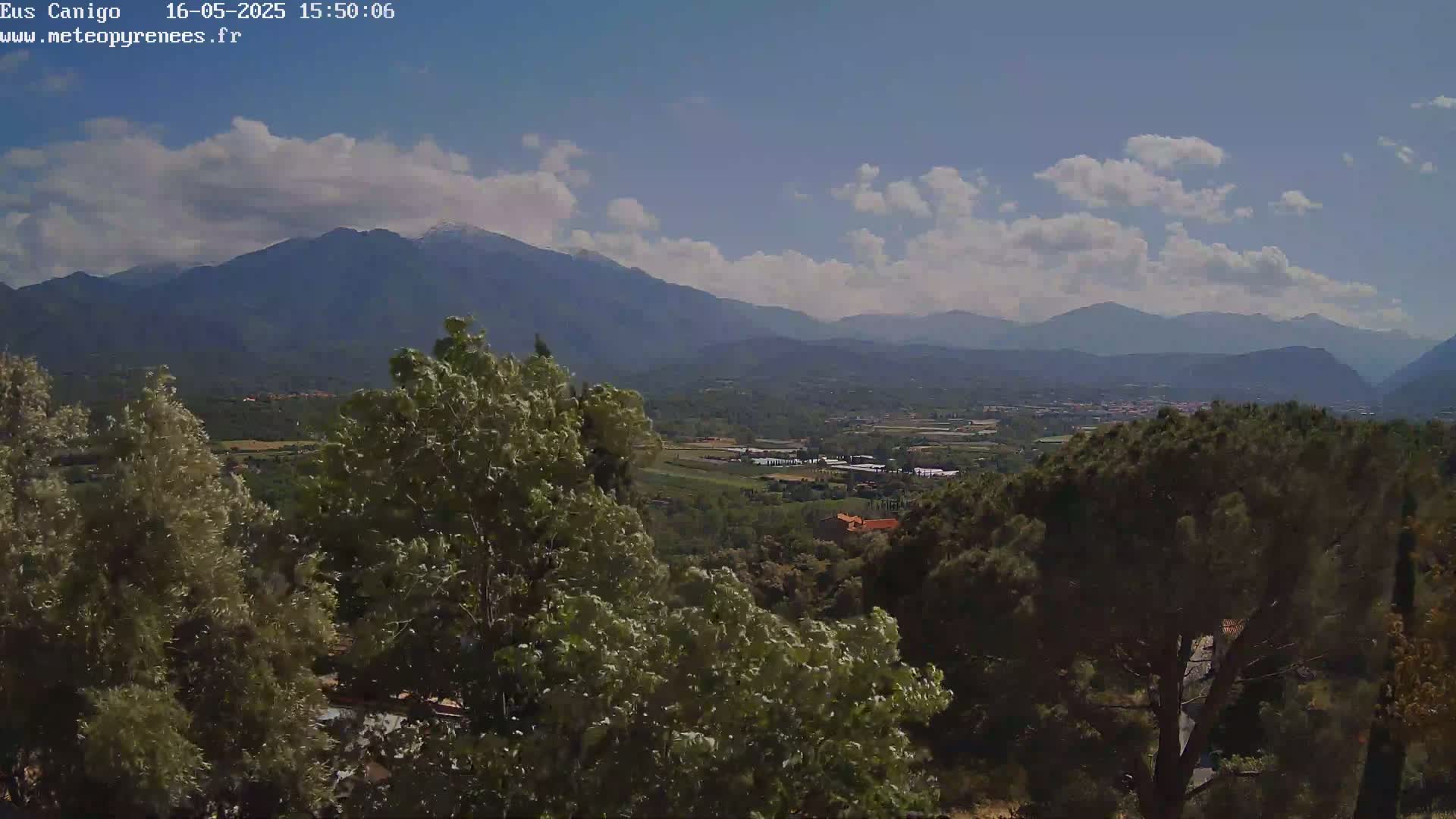 A partly cloudy, sunny day reveals a panoramic view of lush green valleys and distant mountains, some with snow-capped peaks, seen from a high vantage point amongst trees.