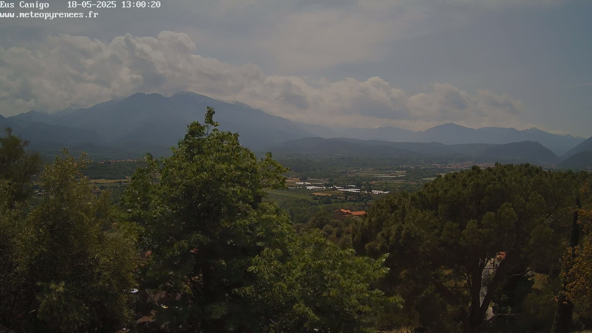 A partly cloudy sky overlooks a valley of green trees and distant mountains, some of which are snow-capped.