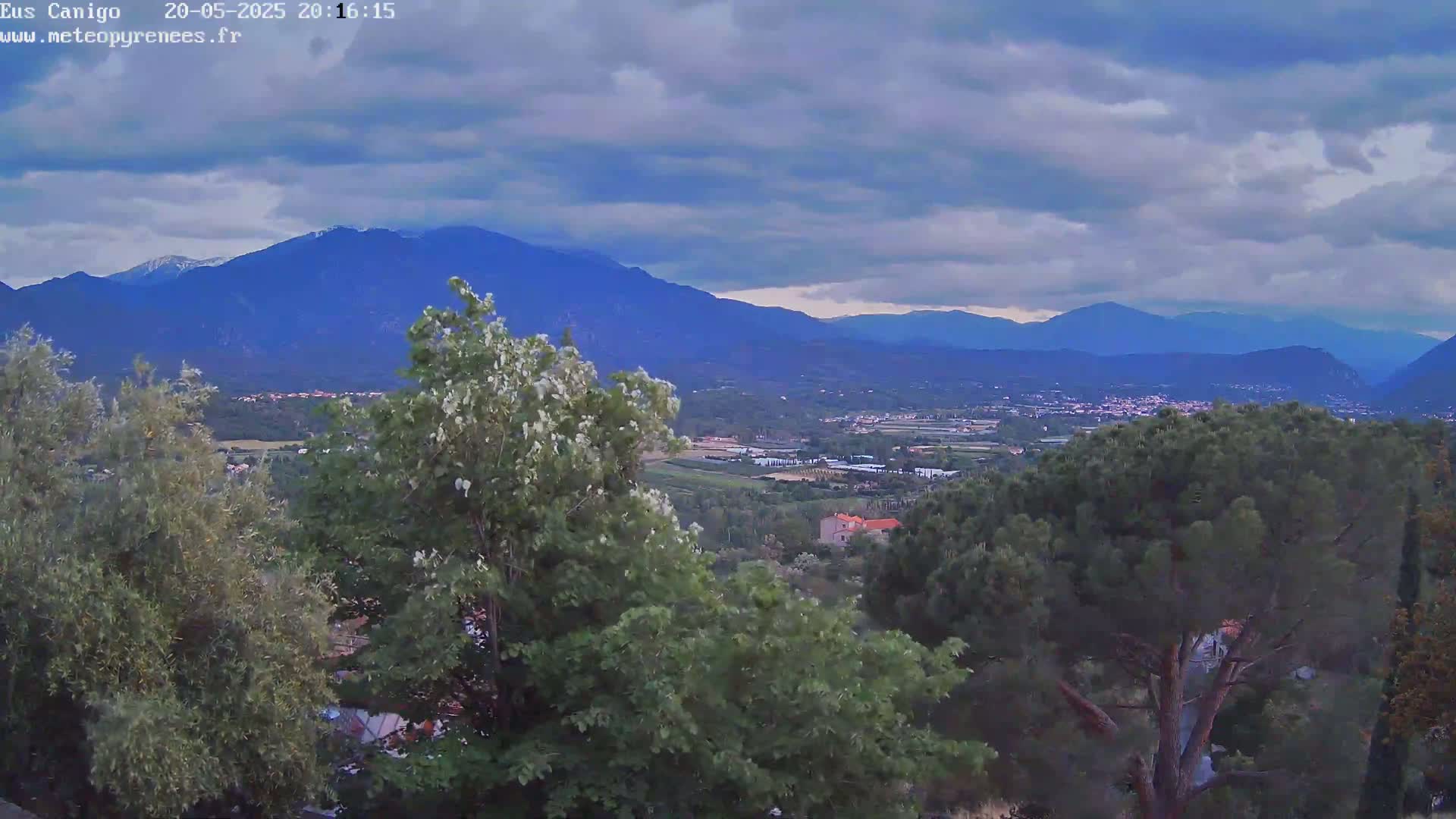 A view from a hilltop shows a valley with trees and buildings under a partly cloudy sky, with mountains in the background.