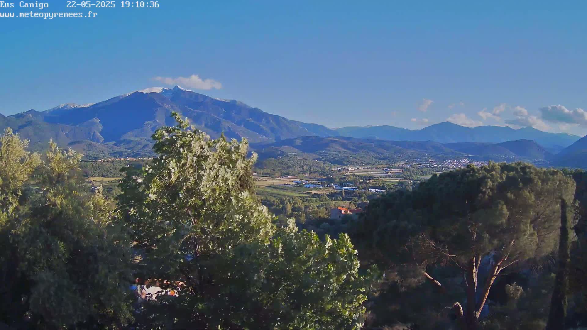A mountain range with a snow-capped peak is visible in the distance, overlooking a valley with trees and a town under a clear, bright sky.