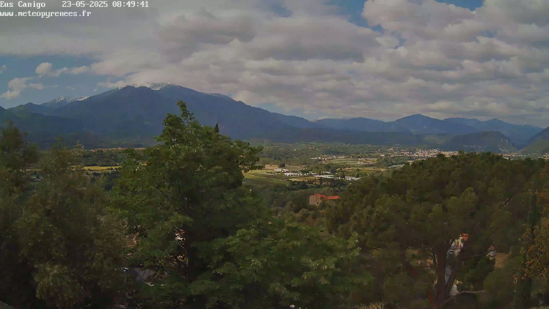 A partly cloudy day reveals a valley with a village nestled between mountains, one of which is snow-capped.