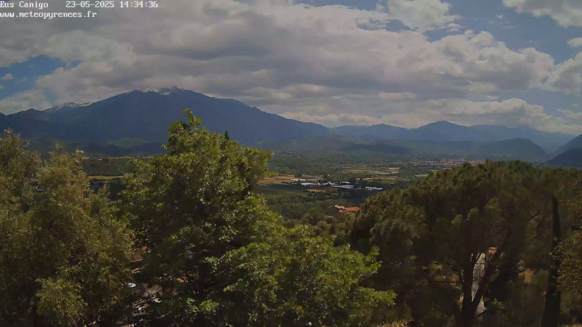 A partly cloudy sky overlooks a valley of green trees and distant mountains, one of which shows hints of snow.