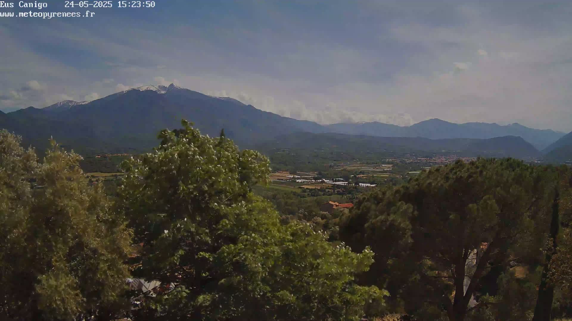 A partly cloudy day reveals a valley view of green trees and distant mountains, one of which has a snow-capped peak.