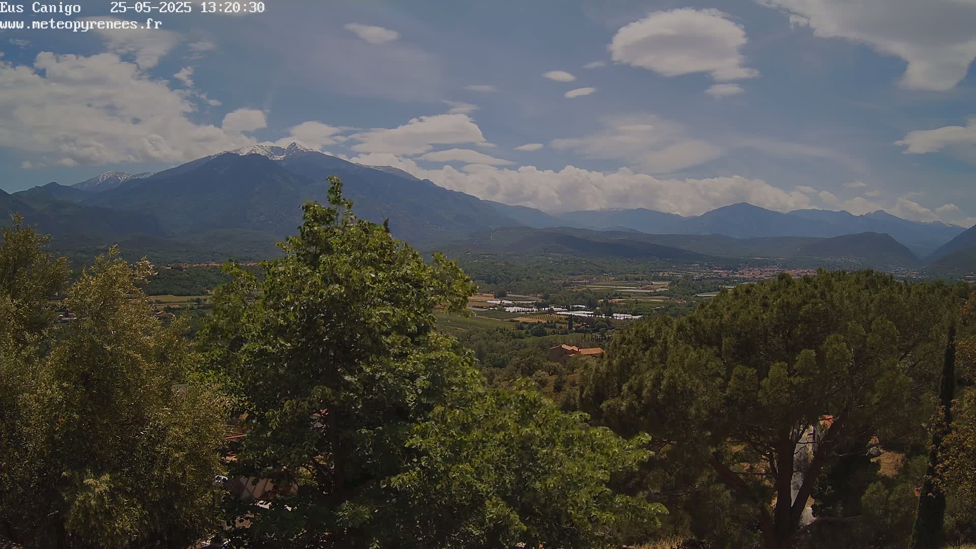 A partly cloudy day reveals a valley view dotted with greenery and buildings, nestled between mountains, one of which has a snow-capped peak.