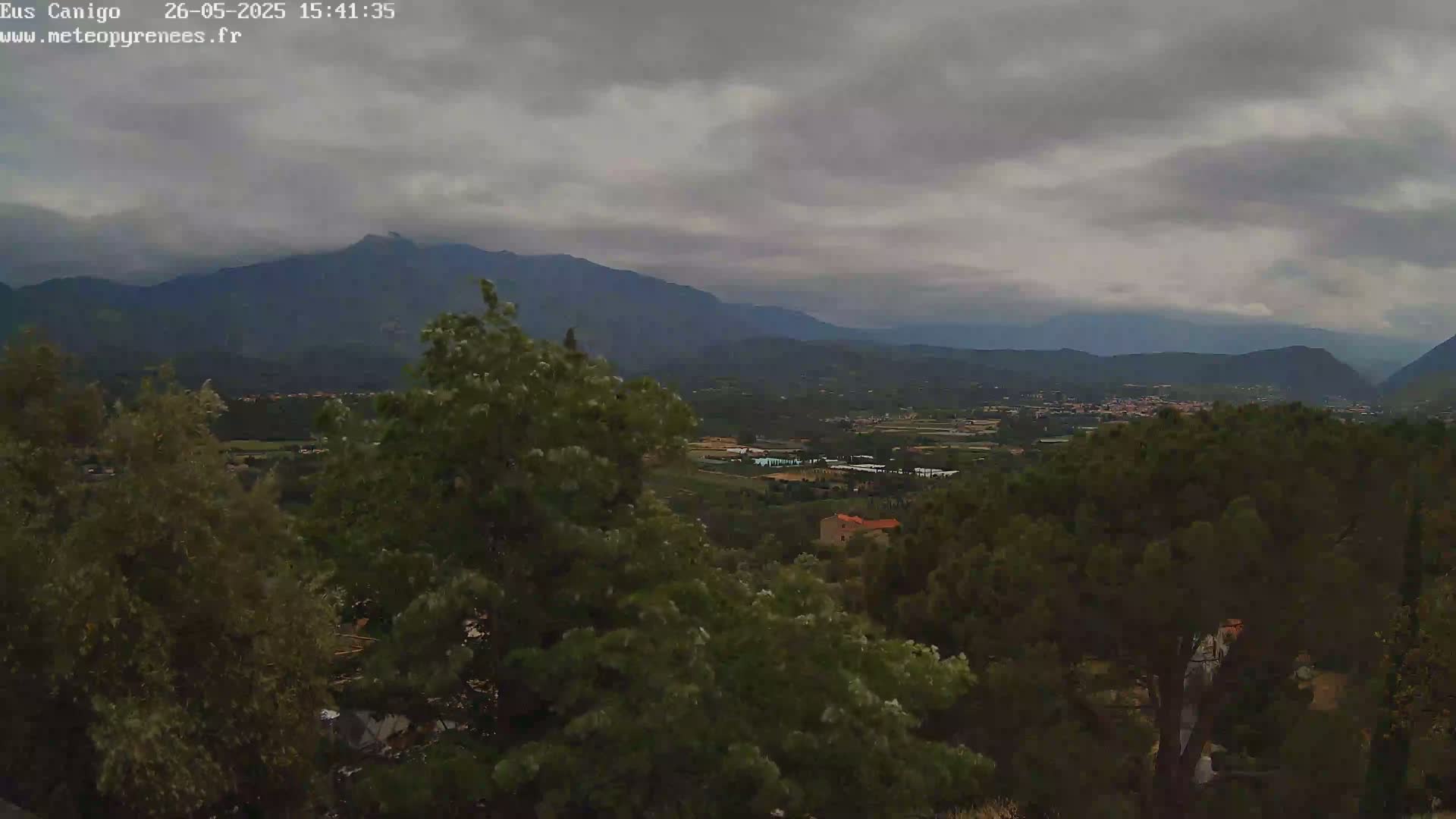 A view of a valley with green trees and a village under an overcast sky, mountains in the distance.