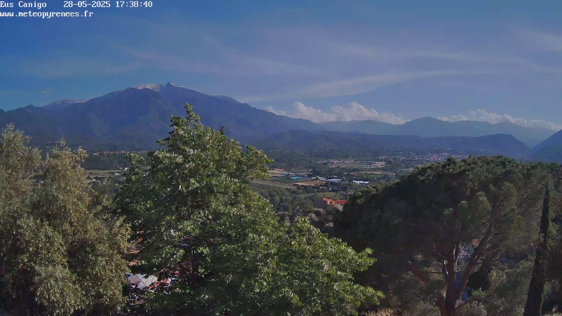 A partly cloudy blue sky overlooks a valley of green trees and distant mountains, one of which is capped with snow.