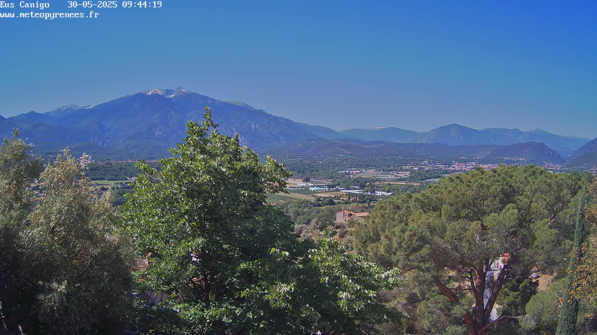 A mountain range with some snow on the peaks is visible in the distance, under a clear blue sky, behind a valley of green trees and farmland.