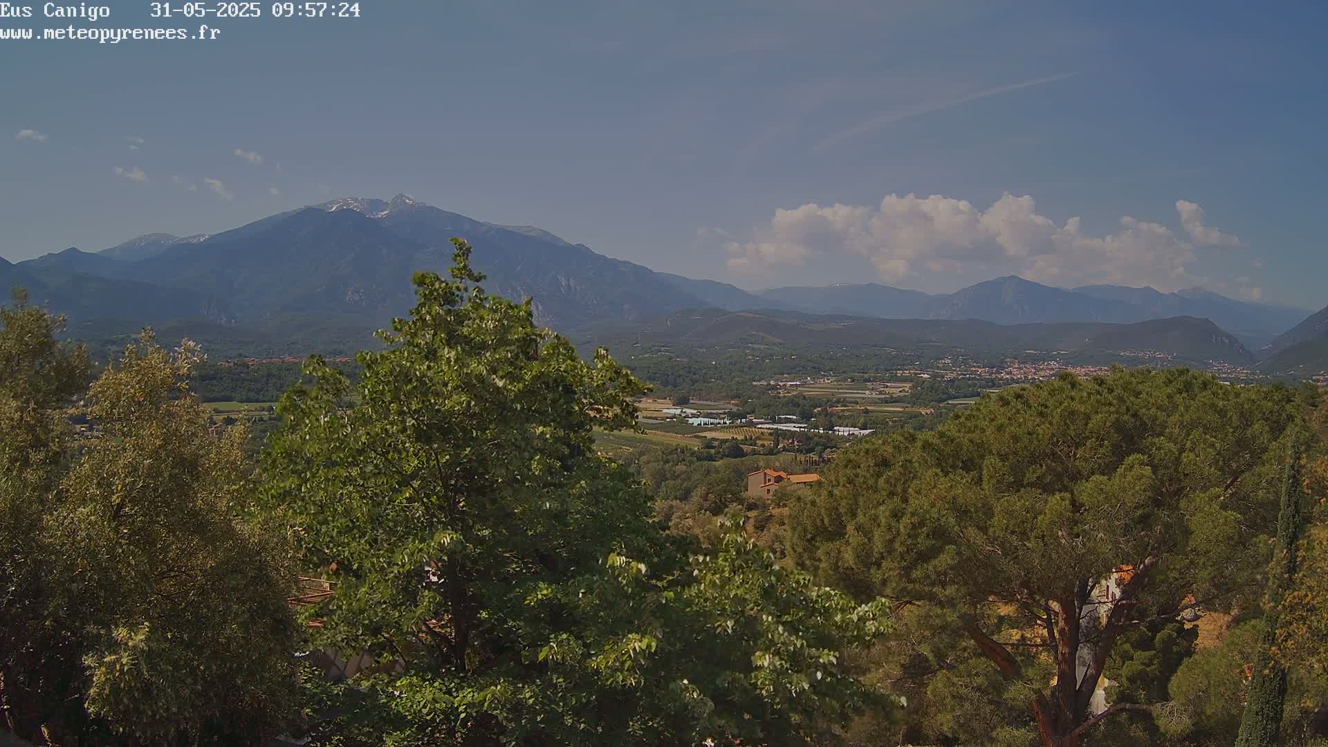 A partly cloudy day reveals a mountainous landscape, with lush green trees in the foreground and a valley stretching towards distant, snow-capped peaks.