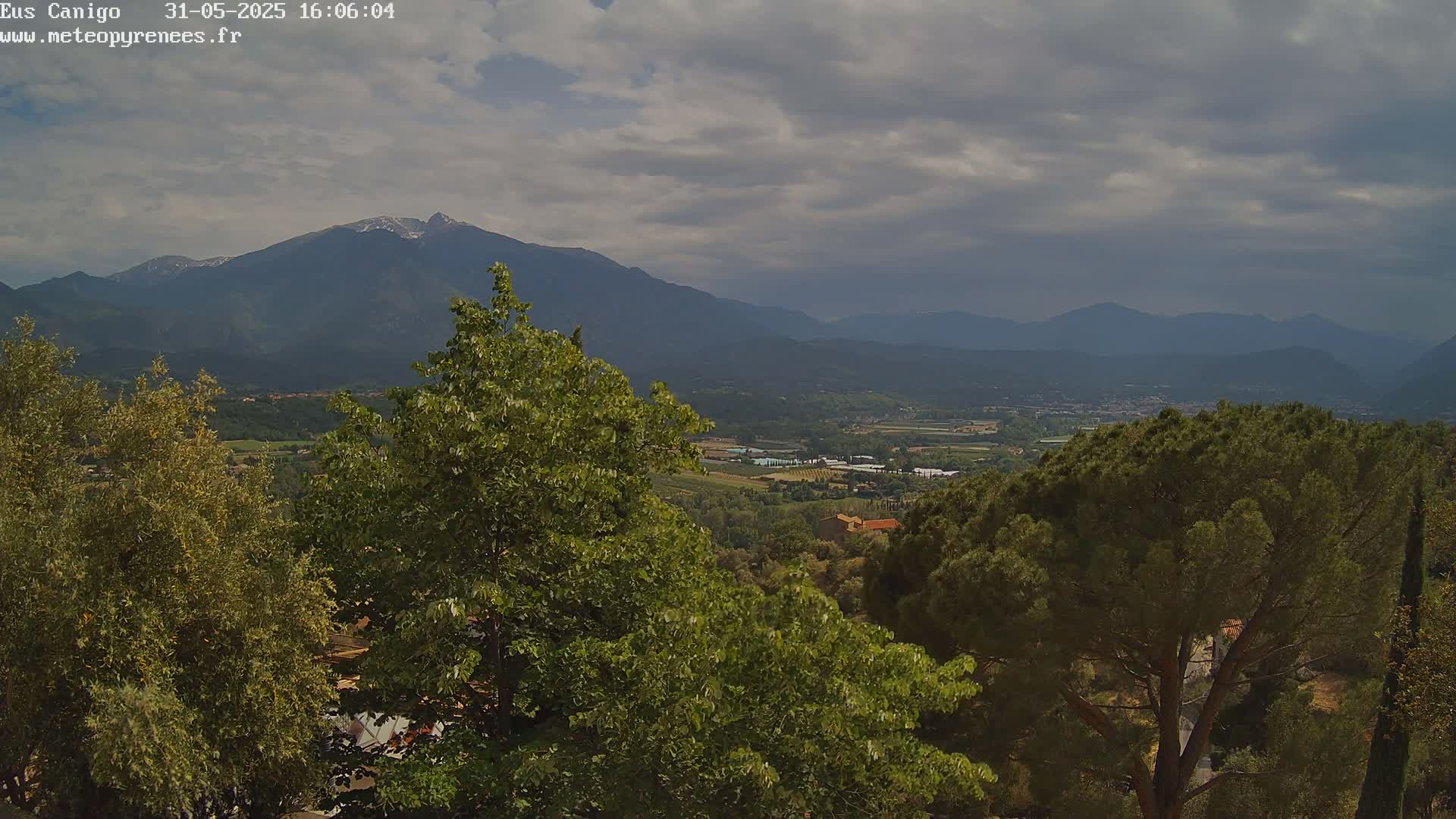 A mountain range with snow-capped peaks is visible in the distance under a cloudy sky, overlooking a valley of green trees and fields.