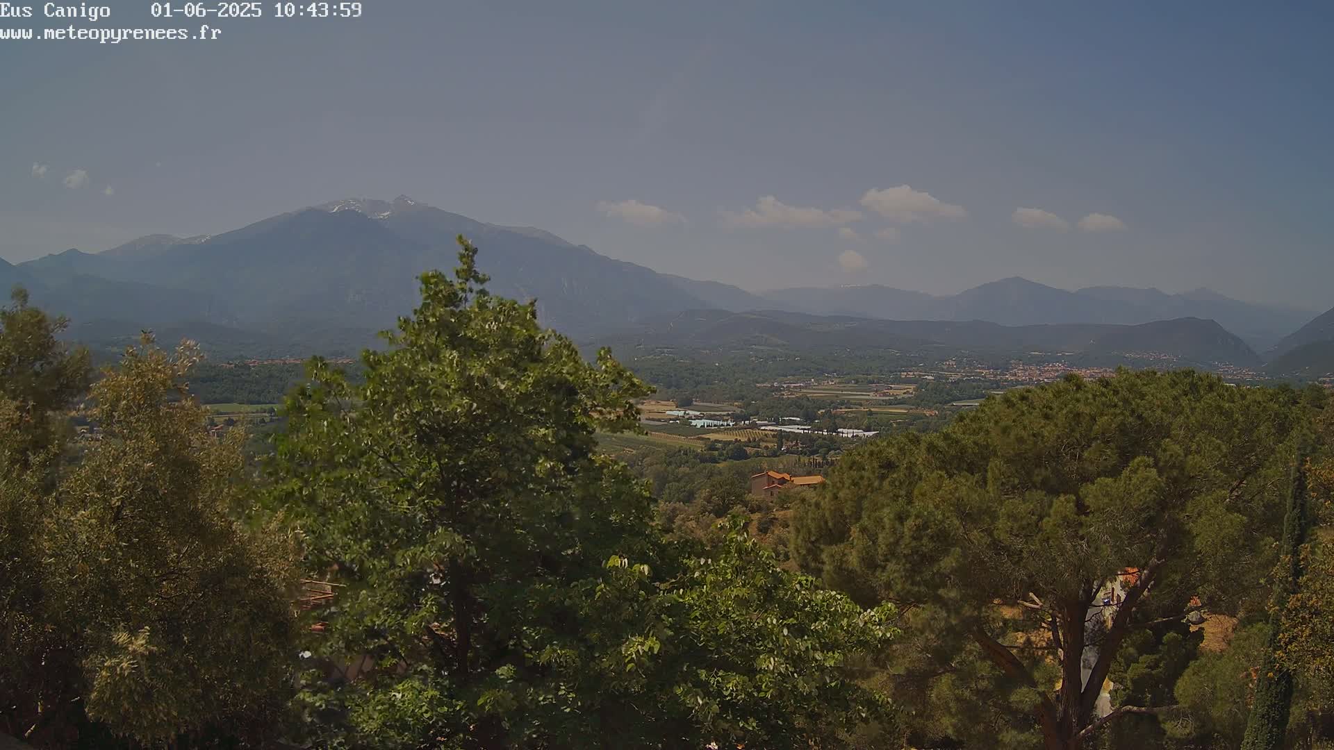 A partly cloudy daytime view of a valley and mountains, with lush green trees in the foreground.