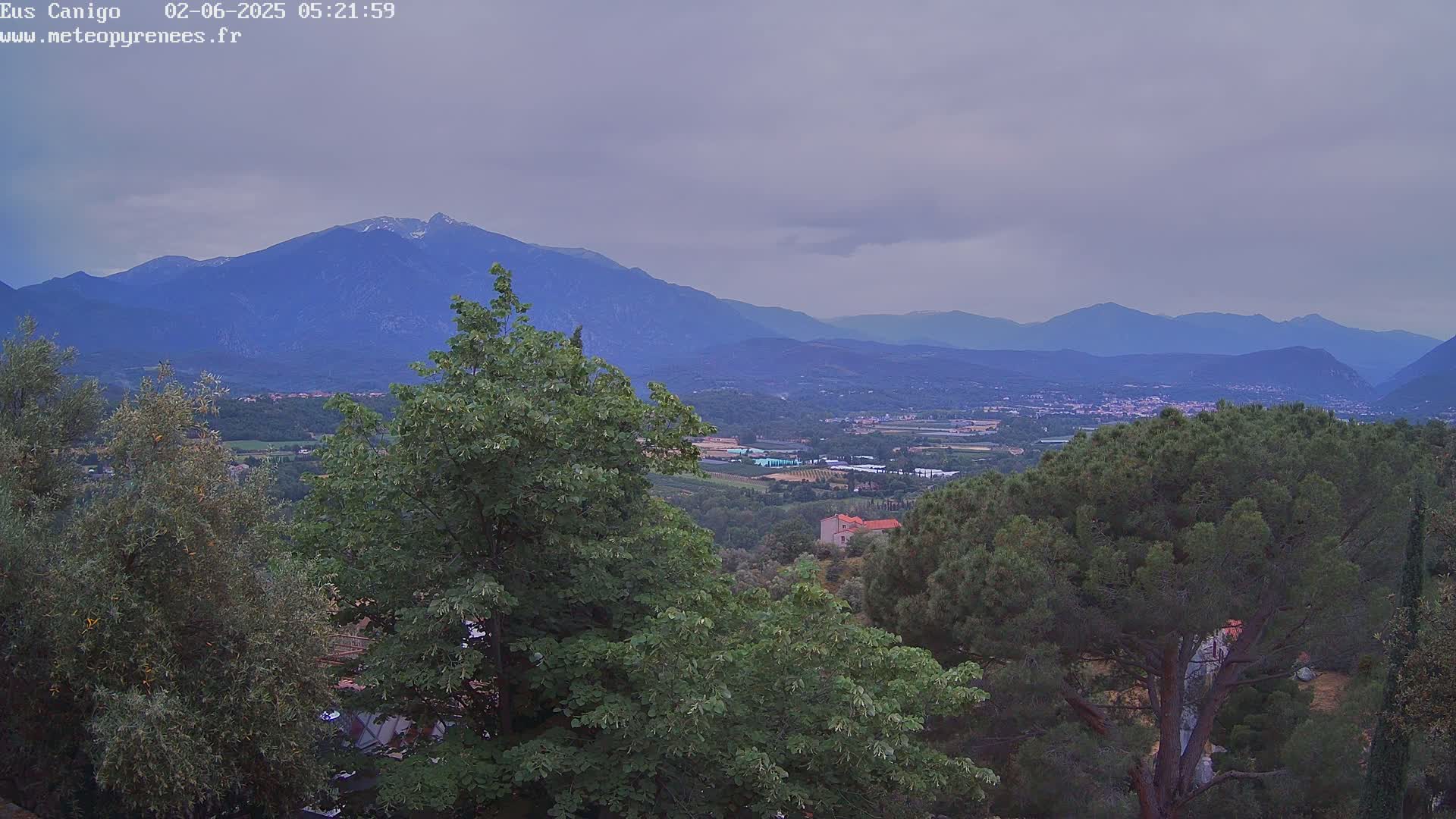 A mountainous landscape under an overcast sky is viewed from an elevated position, showing a valley with trees and buildings in the foreground.