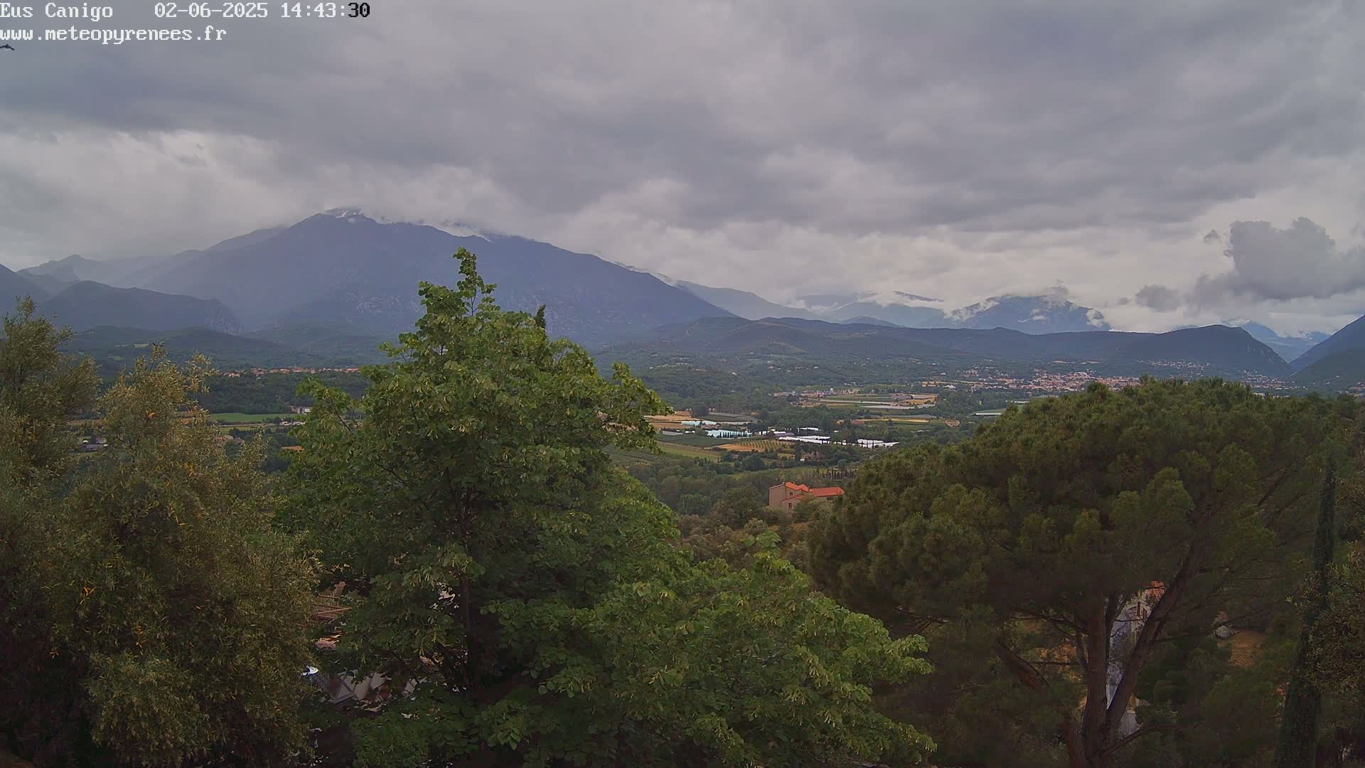 A mountainous landscape under a cloudy sky is viewed from a hilltop, showing lush green trees in the foreground and a valley with a town nestled between distant mountains.