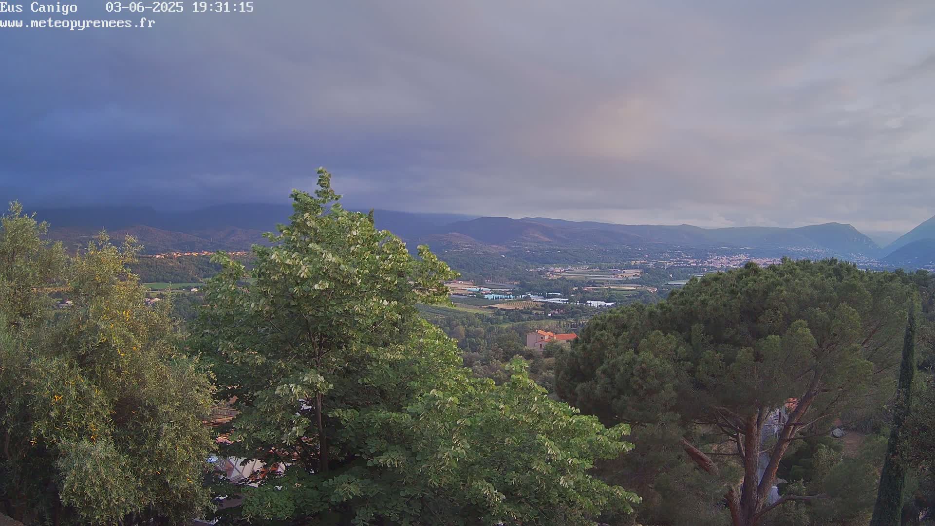 An expansive view of a mountainous landscape under a mostly cloudy sky, showcasing lush green trees in the foreground and a valley with scattered buildings in the distance.